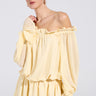 A person models the "ON YOUR MIND CHIFFON MINI DRESS," a pastel yellow, off-the-shoulder dress with ruffled edges and long sleeves, against a white backdrop. The person has blonde hair pulled back and accessorizes with hoop earrings.