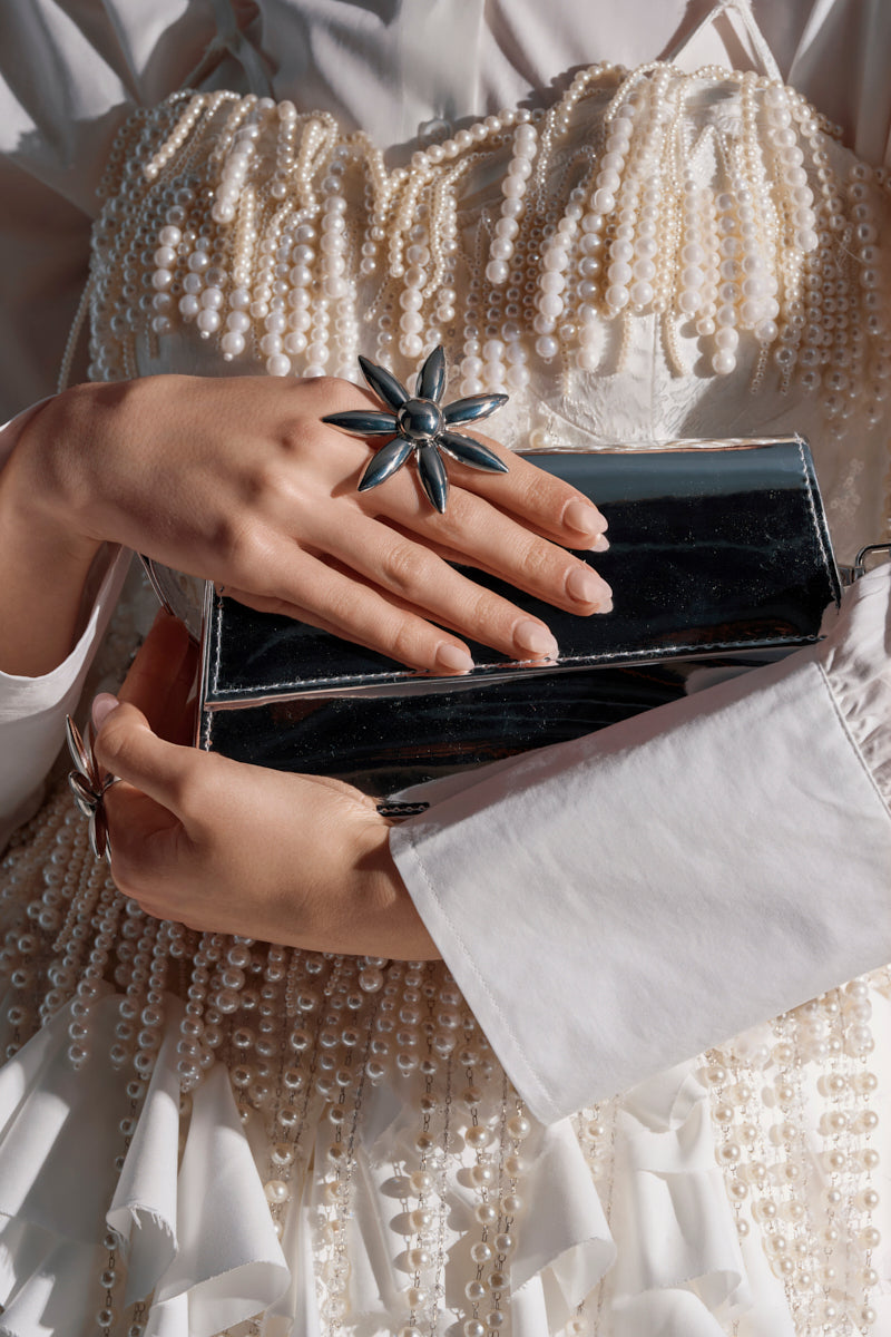 A person in a white pearl-embellished dress holds a shiny silver clutch, showing off manicured nails and the IN THE SUN RING SET—a blooming flower pendant-inspired statement ring with adjustable bands.