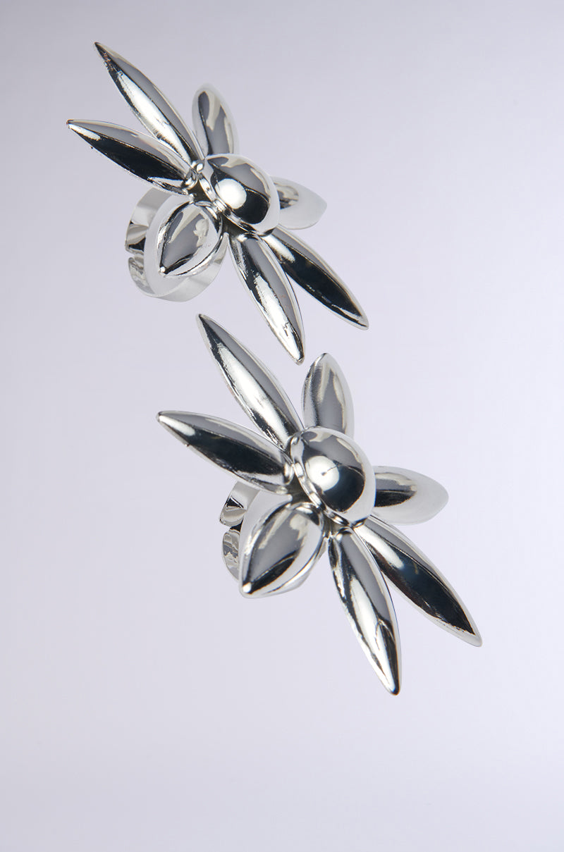 The IN THE SUN RING SET features two shiny silver rings from AKIRA Label, each with six-pointed petal designs resembling flowers, displayed against a plain light background—a stunning accessory for any occasion.