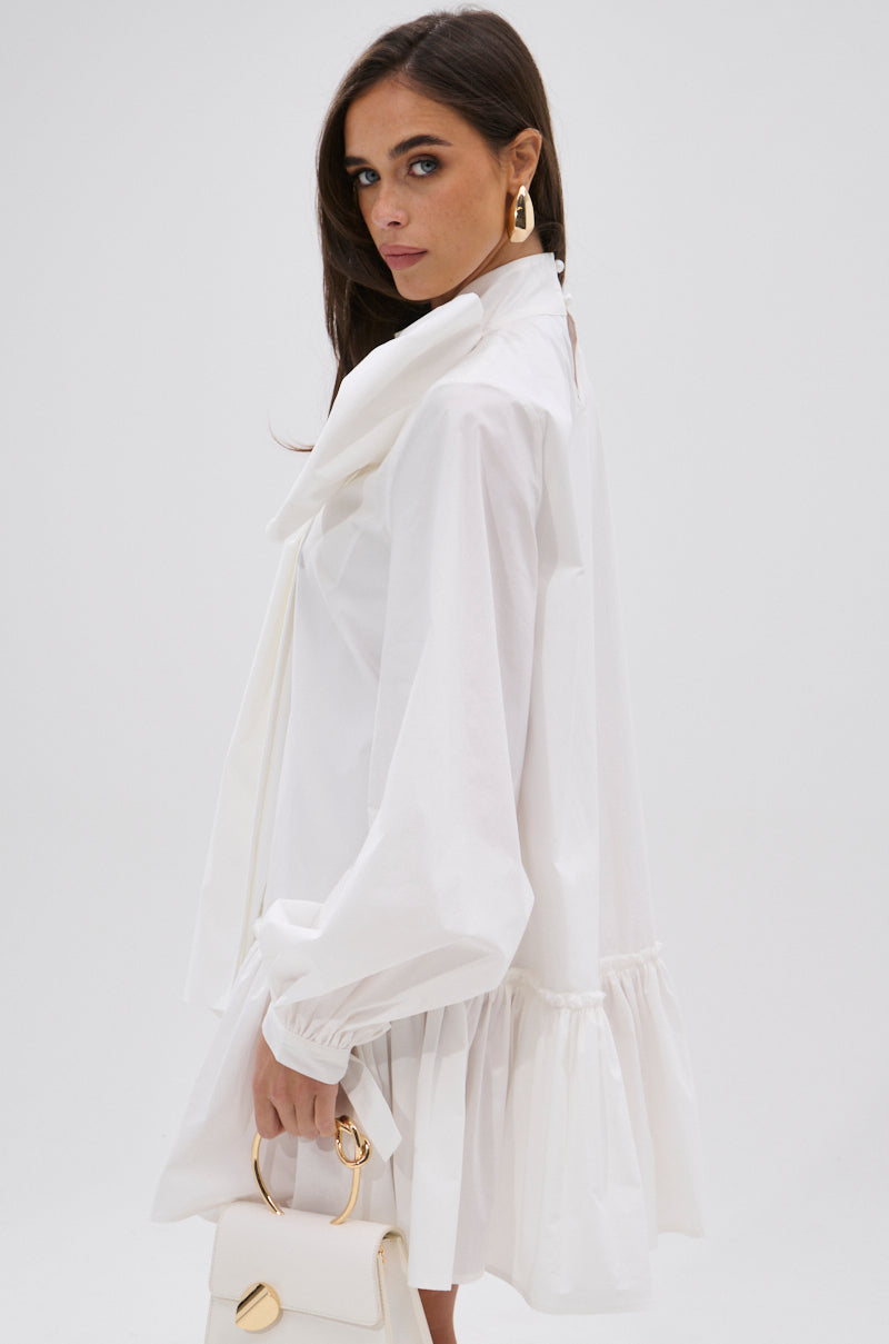 A woman with long brown hair and gold hoop earrings stands sideways against a plain background, holding a white handbag. She wears the loose-fitting TAKE A BOW POPLIN MINI DRESS in white, featuring puff sleeves and a ruffled hem.