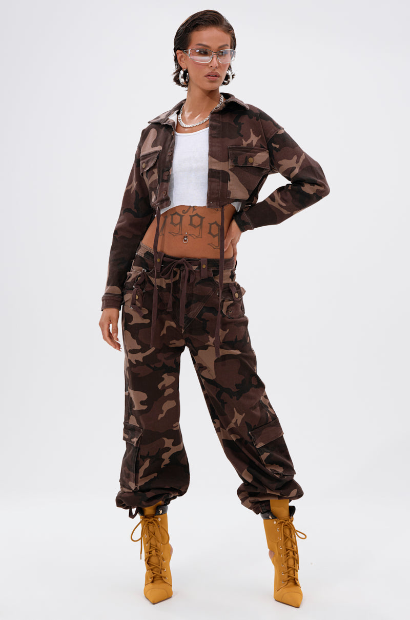 A person poses confidently in the CANT SEE ME BROWN CAMP CARGO JOGGER paired with a cropped camo jacket, white ribbed crop top, clear glasses, hoop earrings, layered necklaces, and mustard-yellow lace-up boots against a plain background.