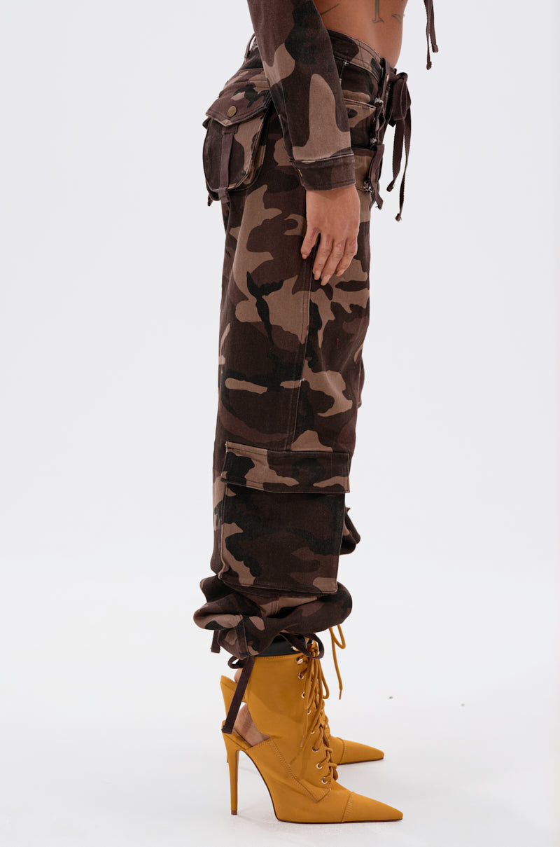 A person stands in profile wearing CANT SEE ME BROWN CAMP CARGO JOGGER pants and mustard yellow pointed-toe lace-up high heels against a plain white background.
