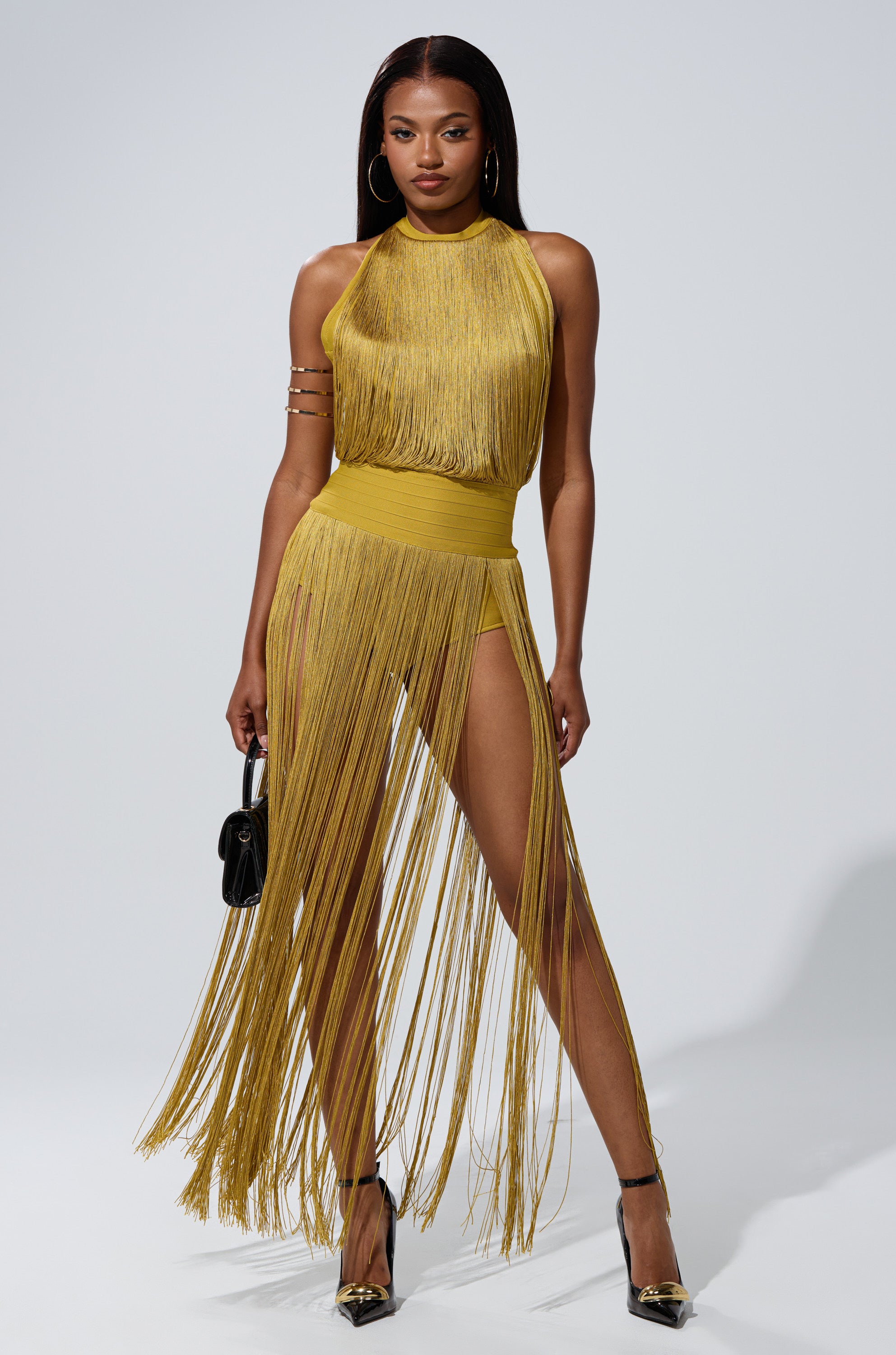 A woman poses confidently in the RENEE FRINGE ROMPER IN GREEN, featuring a high waist and long fringe panels. She holds a small black handbag, wears gold heels and hoop earrings, standing against a plain background.
