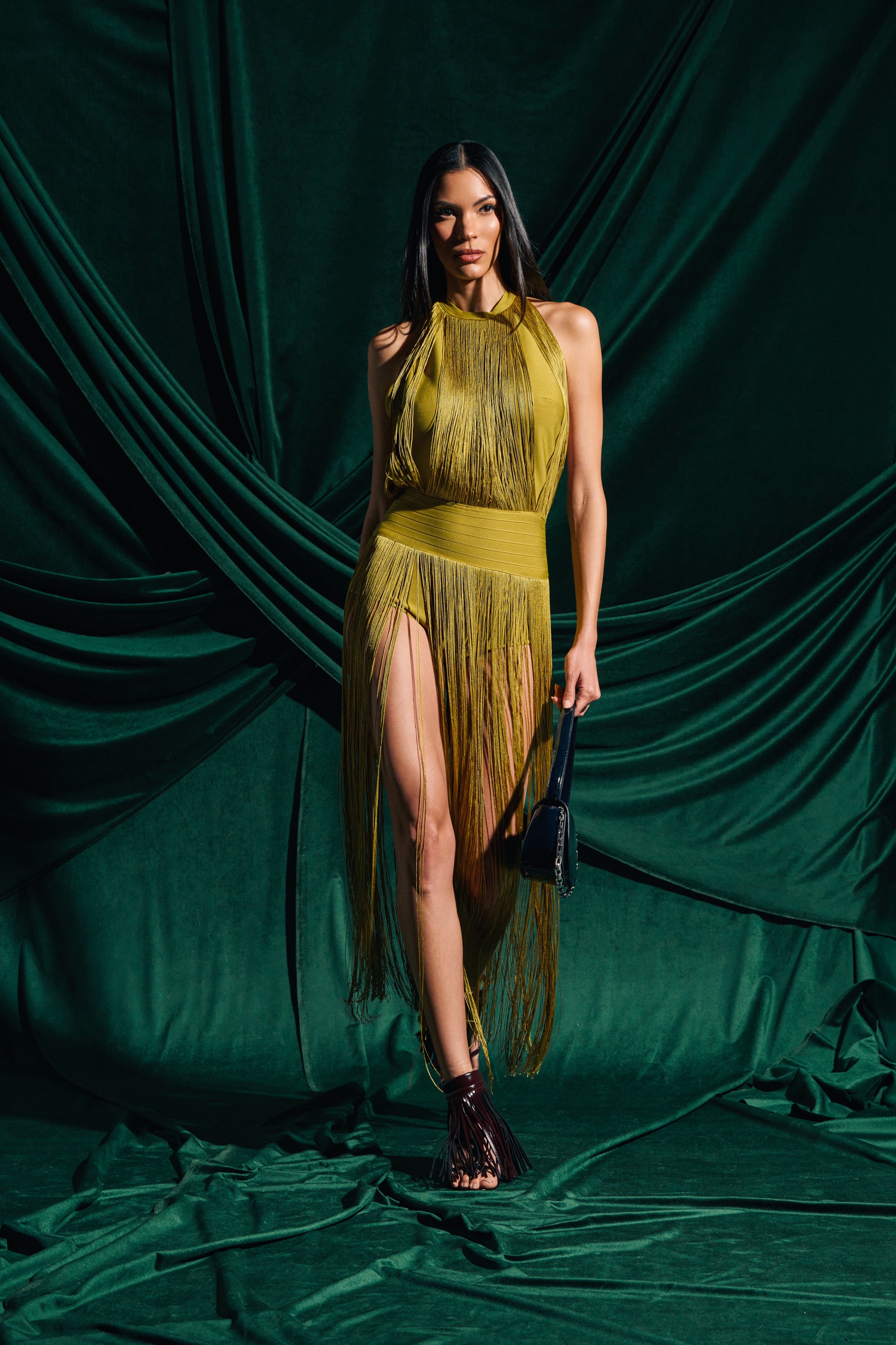 A woman poses against a dark green backdrop in the RENEE FRINGE ROMPER IN GREEN, featuring a sleeveless design, yellow-green fringe, thigh-high slit, and alluring open back. She accessorizes with a small black handbag and black strappy shoes.