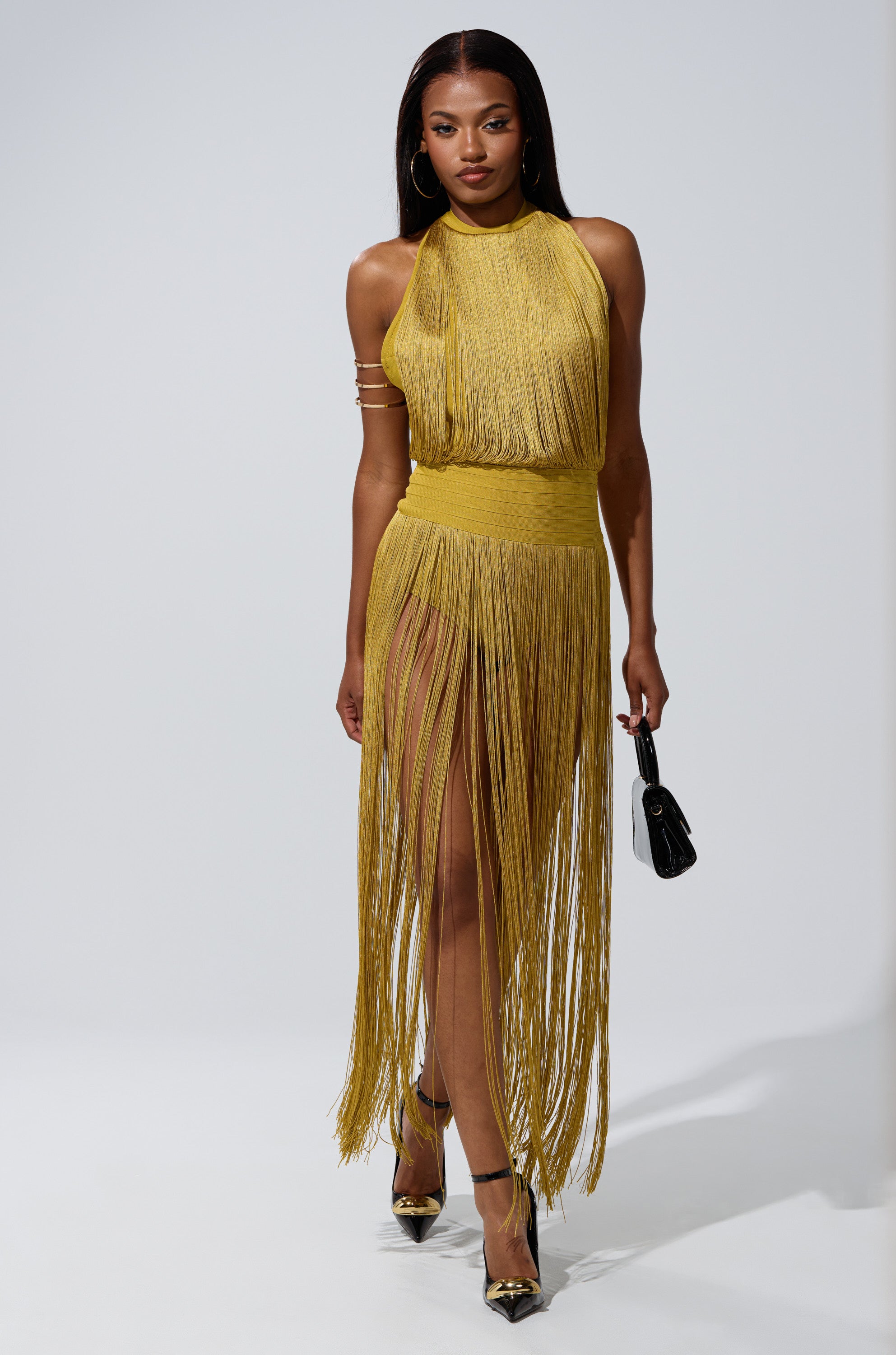 A woman poses confidently against a plain white background wearing the RENEE FRINGE ROMPER IN GREEN, styled with black heels and a small black handbag.