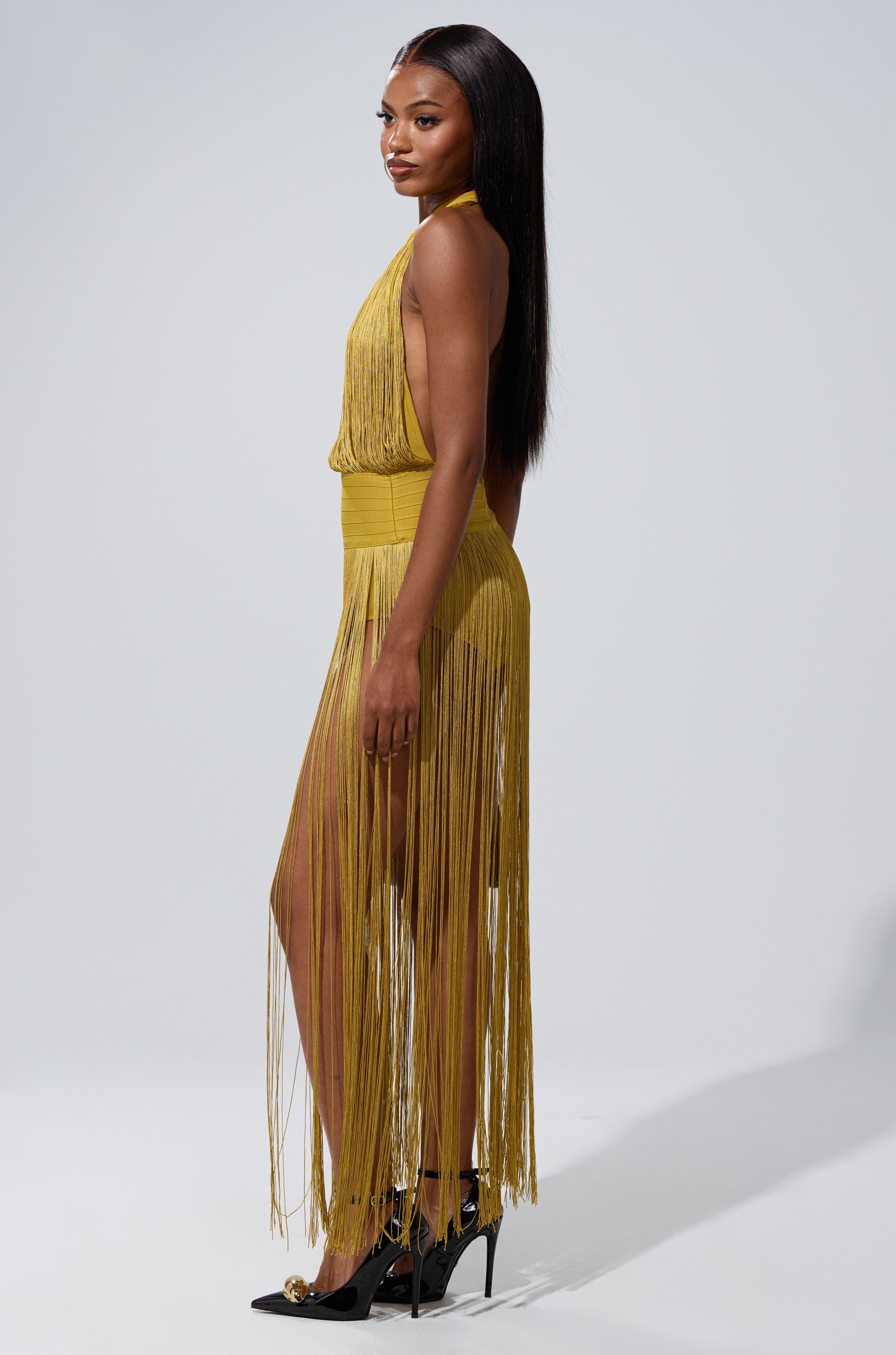 A woman stands in profile, exuding confidence in the RENEE FRINGE ROMPER IN GREEN paired with black heels. Her long, straight hair flows as she poses against a plain gray background.