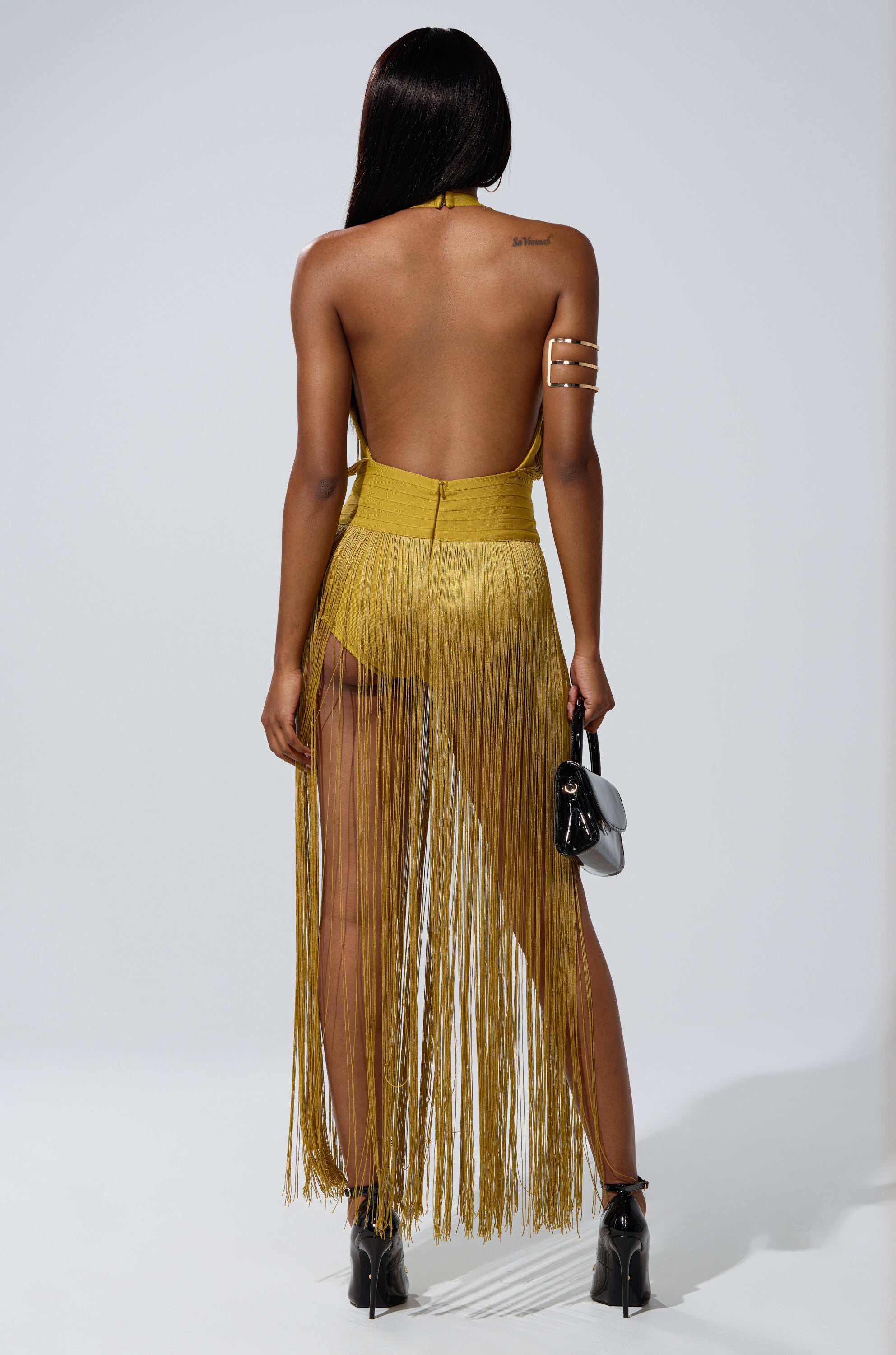 A woman faces away, wearing the RENEE FRINGE ROMPER IN GREEN with long fringe, black high-heeled ankle boots, gold arm cuffs, and holding a small black handbag. Her straight hair flows down her back.