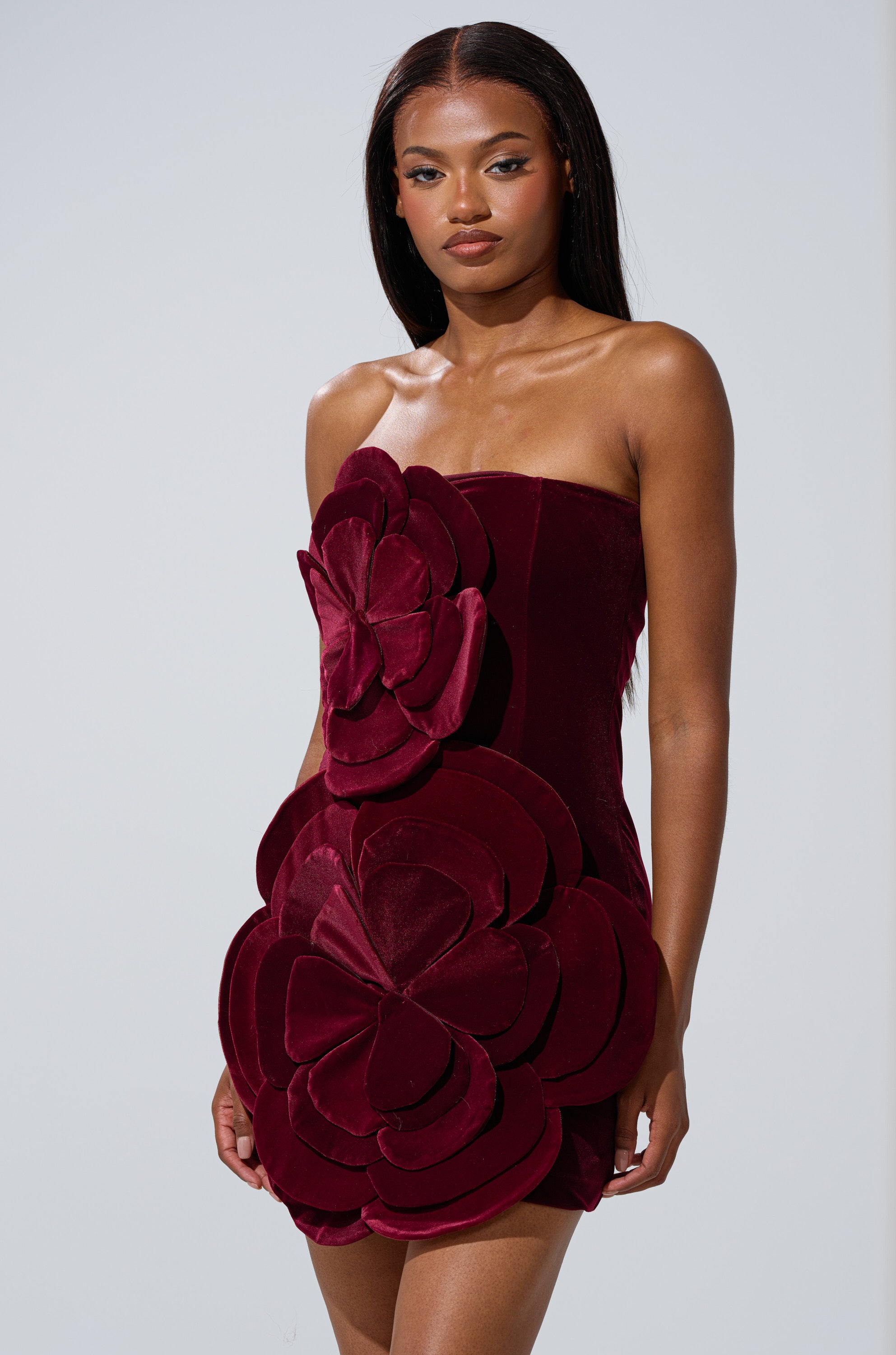 A woman stands against a light gray background wearing the FLOWER POWER VELVET MINI DRESS, a strapless mini dress featuring large 3D floral appliqués on the bodice and skirt.