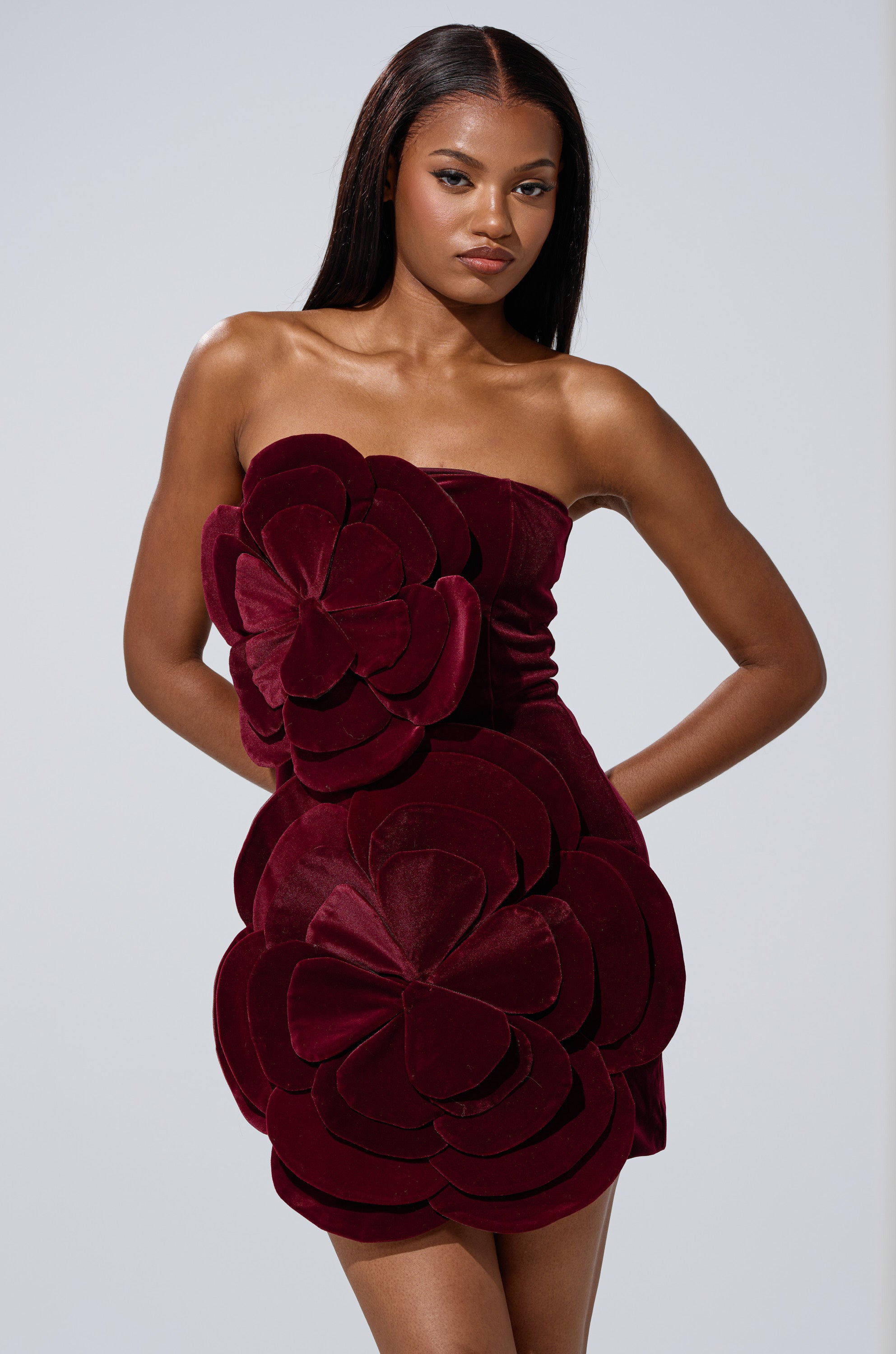 A woman models the FLOWER POWER VELVET MINI DRESS, featuring a strapless silhouette and bold 3D floral appliqué on the bodice and skirt, against a plain light gray background.