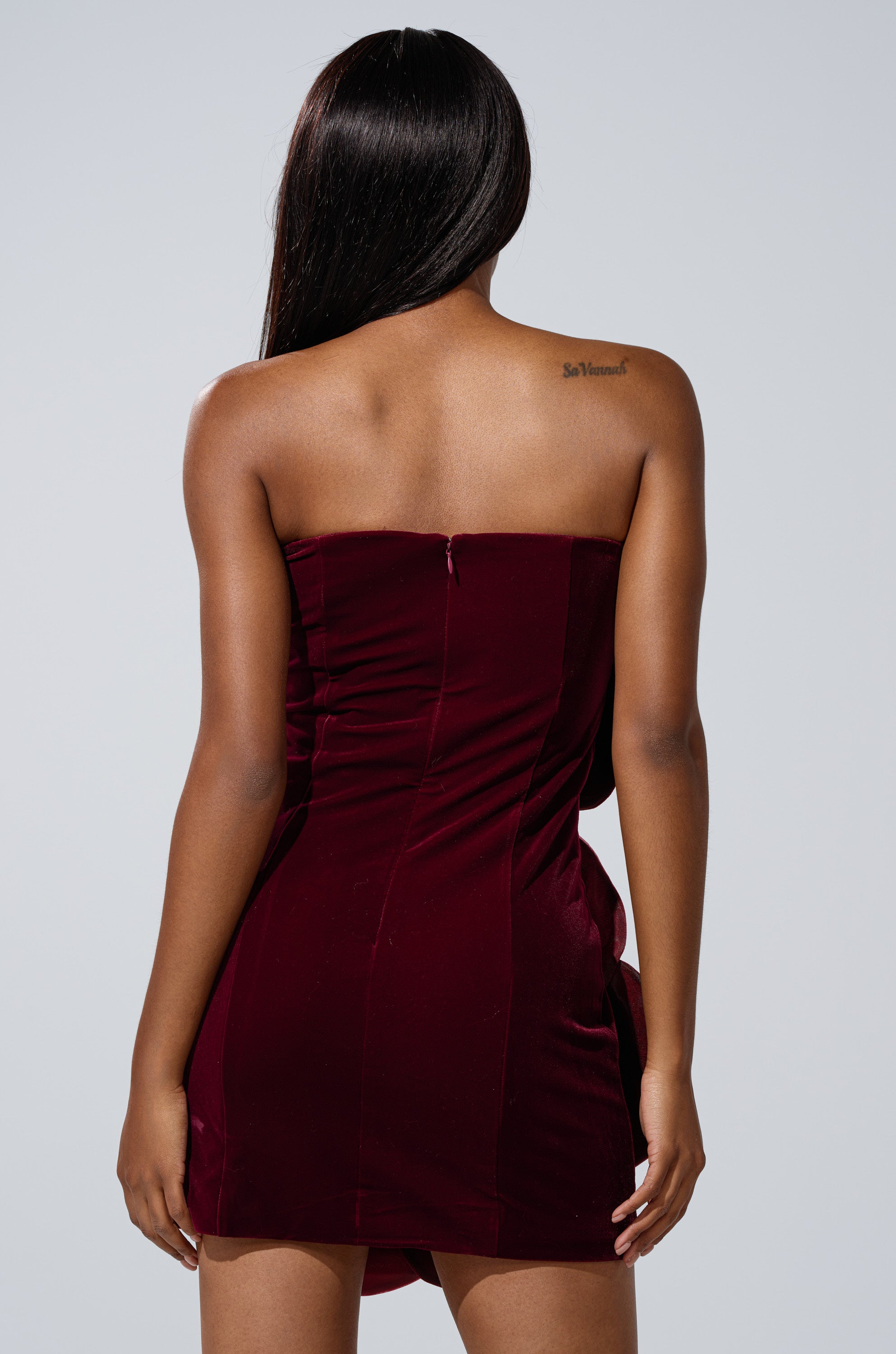 A woman with long, straight dark hair models the FLOWER POWER VELVET MINI DRESS in burgundy with a strapless neckline, her back to the camera showing a tattoo on her upper right shoulder, set against a plain light gray background.