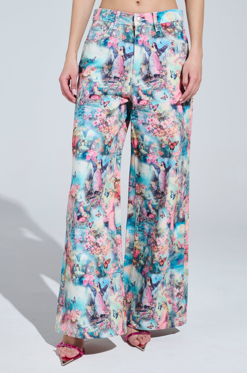 Against a plain background, someone wears FEMALE FANTASY PRINTED DENIM PANTS with a vibrant candy-colored abstract floral pattern in pink, blue, and green hues, paired with pink sandals.