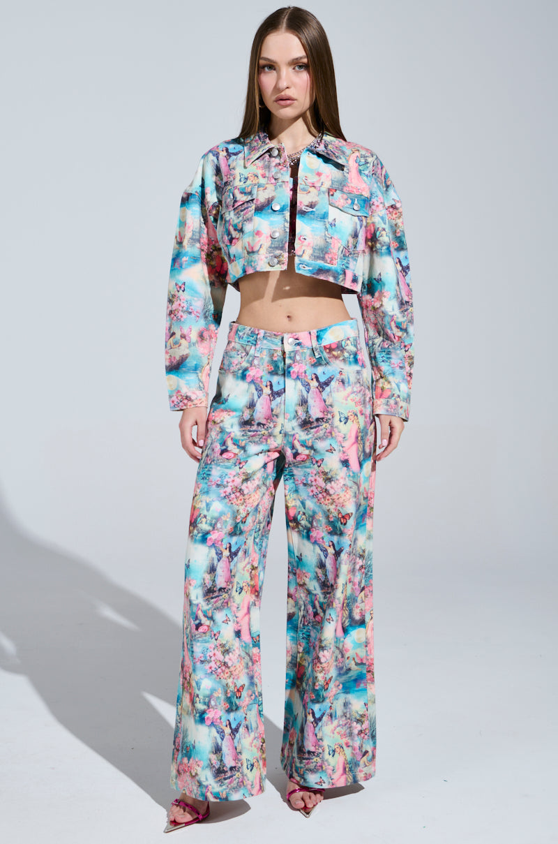 A person stands in a candy-colored print outfit with floral and scenic designs, featuring a cropped jacket and wide-leg FEMALE FANTASY PRINTED DENIM PANTS. The plain background accentuates the vibrant attire.