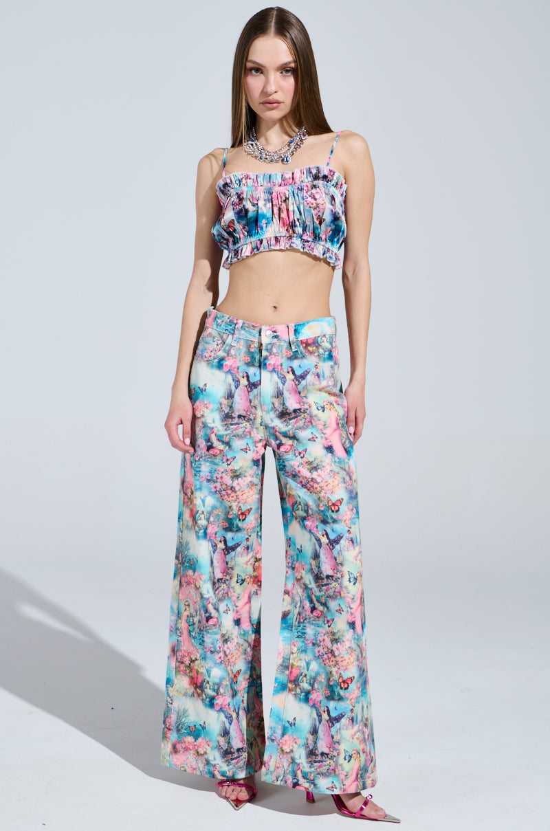 Against a neutral backdrop, a woman showcases the FEMALE FANTASY PRINTED DENIM PANTS in a vibrant floral ensemble. Her sleeveless, ruffled crop top pairs with wide-leg pants, while her long hair and pink pointed-toe heels complete the chic look.