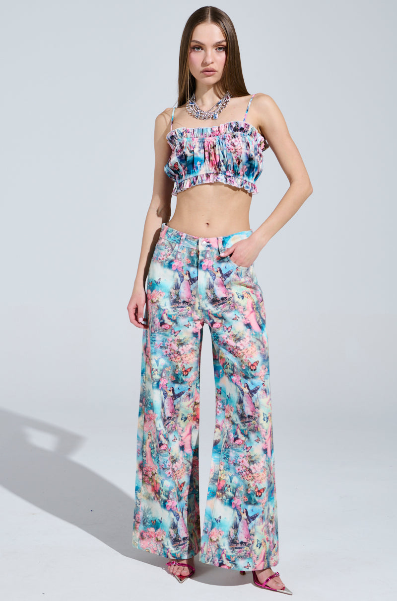 A woman confidently poses in the FEMALE FANTASY PRINTED DENIM PANTS, a candy-colored floral two-piece featuring a ruffled crop top and wide-leg pants. She stands against a plain background, hands in her pockets, wearing pink heels.