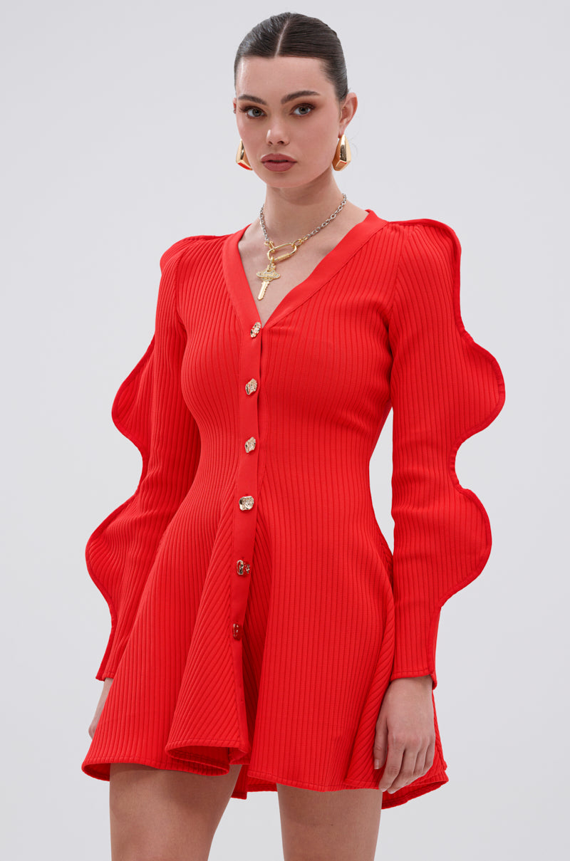 A woman models the COOLEST ON THE BLOCK MINI DRESS IN RED—featuring a bright red ribbed knit, ripple sleeves, and gold buttons—against a light gray background. She pairs it with gold earrings, a necklace, and wears a confident expression.
