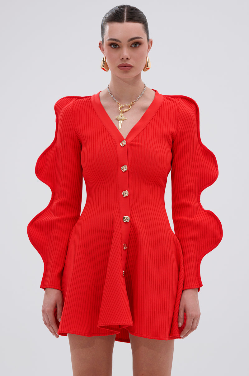 Wearing the COOLEST ON THE BLOCK MINI DRESS IN RED, a woman shows off its vibrant ribbed knit, sculpted ripple sleeves, and gold front buttons. She completes the look with gold earrings and a chain necklace against a light gray background.