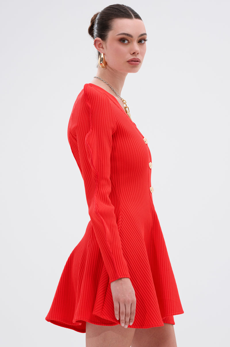 A woman stands in profile against a plain background, wearing the COOLEST ON THE BLOCK MINI DRESS IN RED—ribbed knit with gold buttons. Her hair is in a sleek bun, accessorized with chunky gold earrings and a necklace.