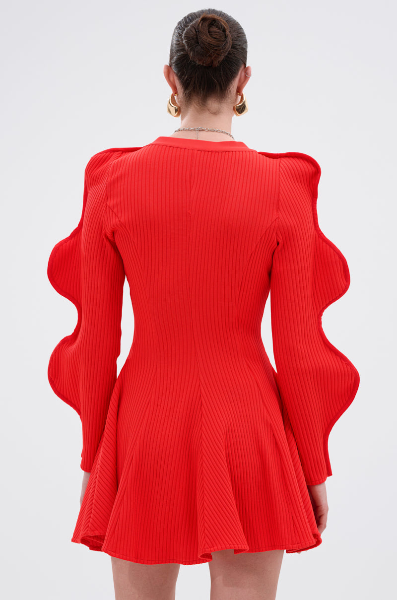 Wearing the COOLEST ON THE BLOCK MINI DRESS IN RED, a woman showcases its ribbed knit fabric, dramatic ripple sleeves, and flared skirt from the back. Her hair is in a neat bun and she pairs the look with large hoop earrings.