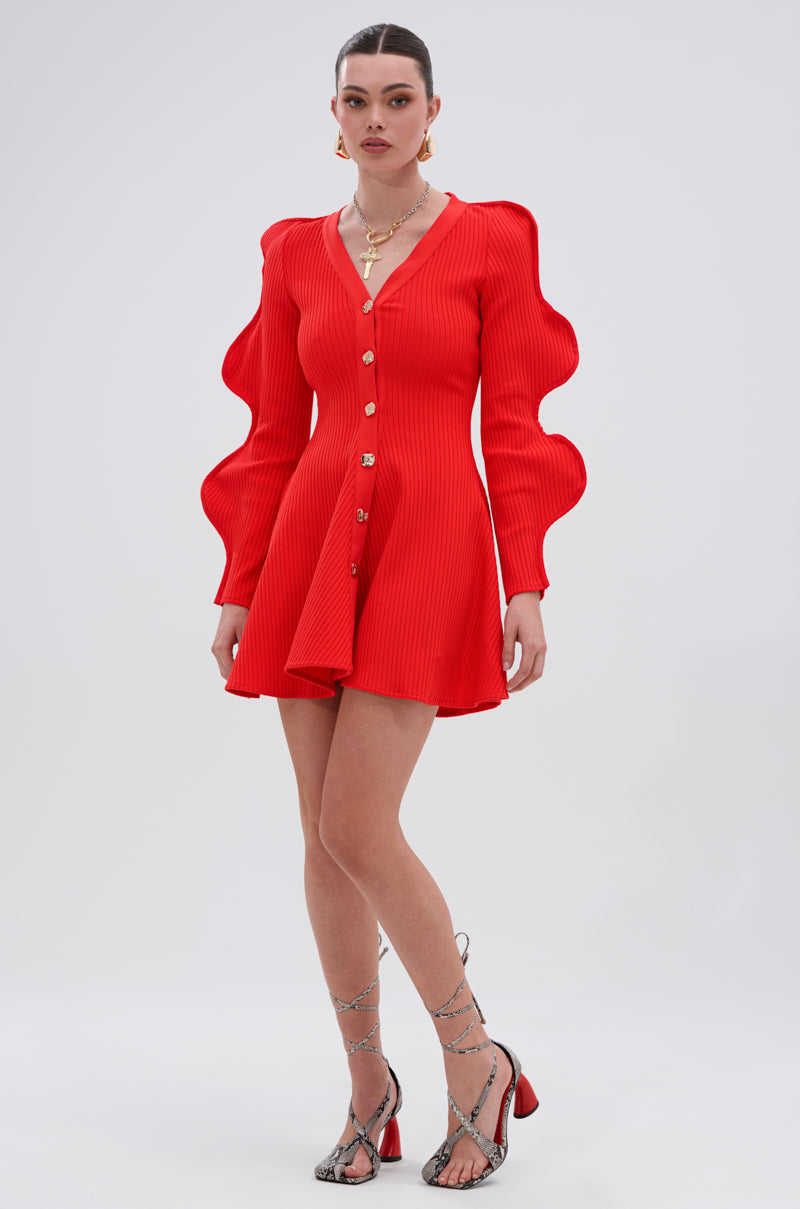 A woman stands against a plain background in the COOLEST ON THE BLOCK MINI DRESS IN RED, featuring dramatic ripple sleeves. She pairs it with silver strappy heels, layered necklaces, and a sleek bun.