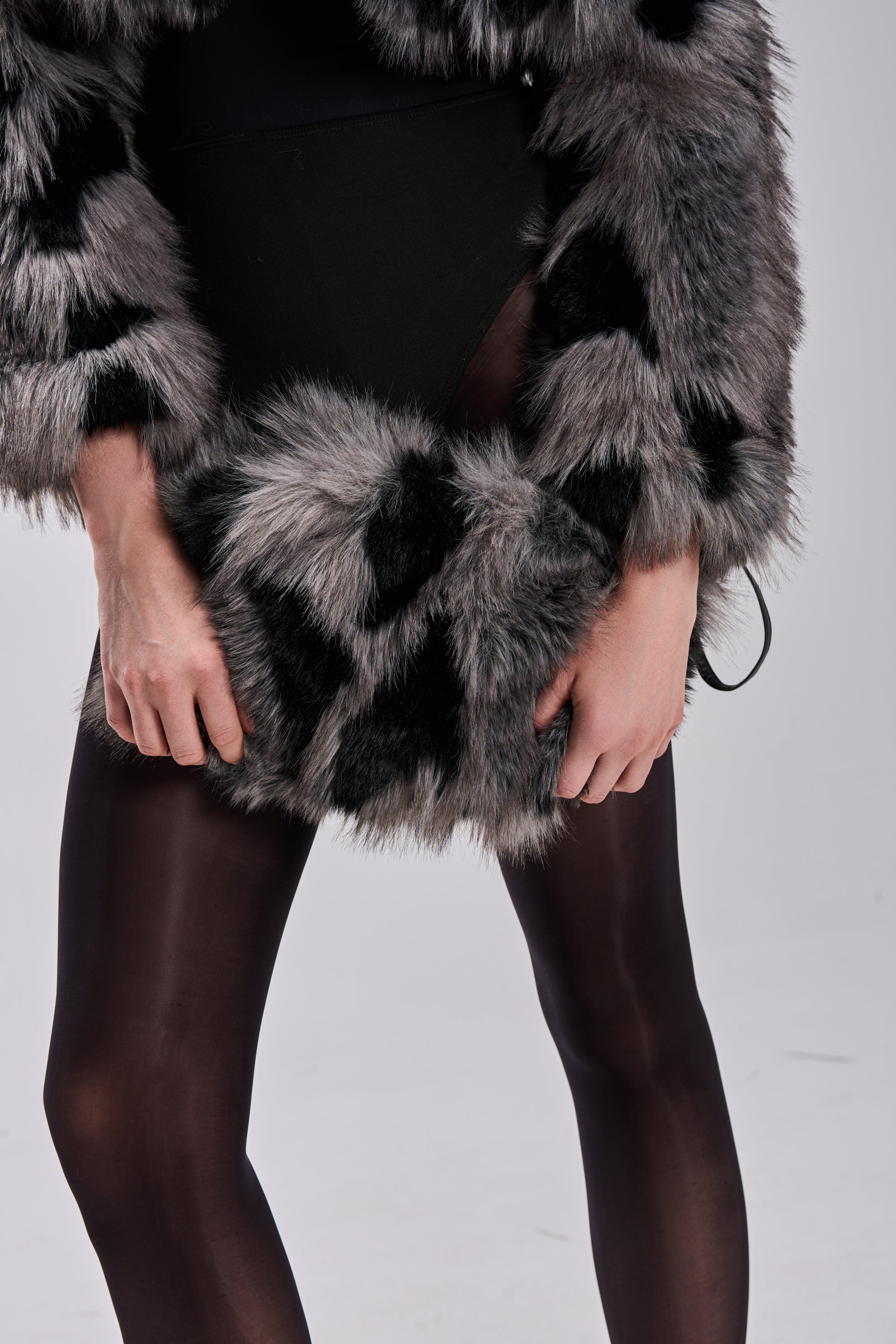 Wearing sheer black tights, a black bodysuit, and a grey and black faux fur coat, a person stands against a light background, holding the ARLO FAUX FUR WRISTLET. The image is cropped to show only the lower torso and upper legs.