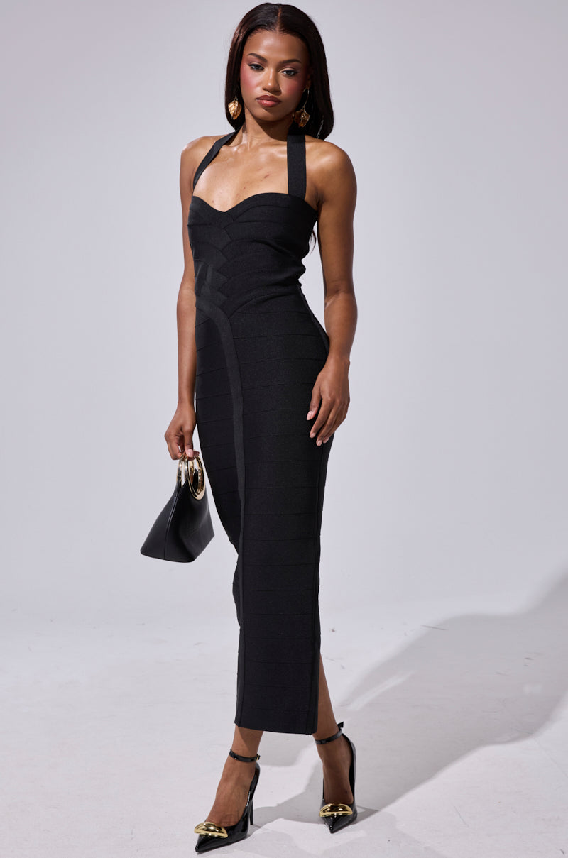 A woman poses against a plain light background wearing the LOVELY DAYS BANDAGE MIDI DRESS, a fitted sleeveless black dress with wide straps. She accessorizes with a black handbag, gold earrings, and black heels with gold accents.