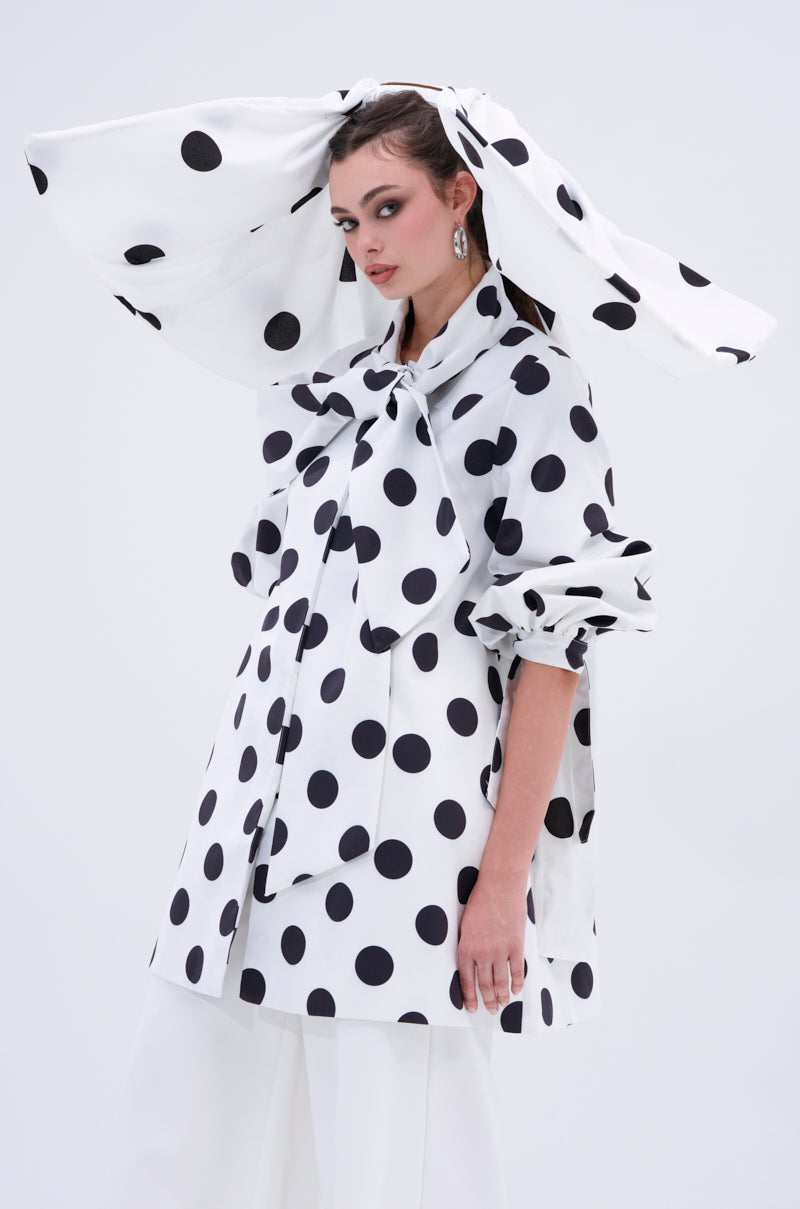 A woman wears the POLKA DOT IT HAIR BOW, a matching oversized accessory with an exaggerated hat and wide sleeves, all in bold white fabric with large black polka dots, standing against a plain light background.