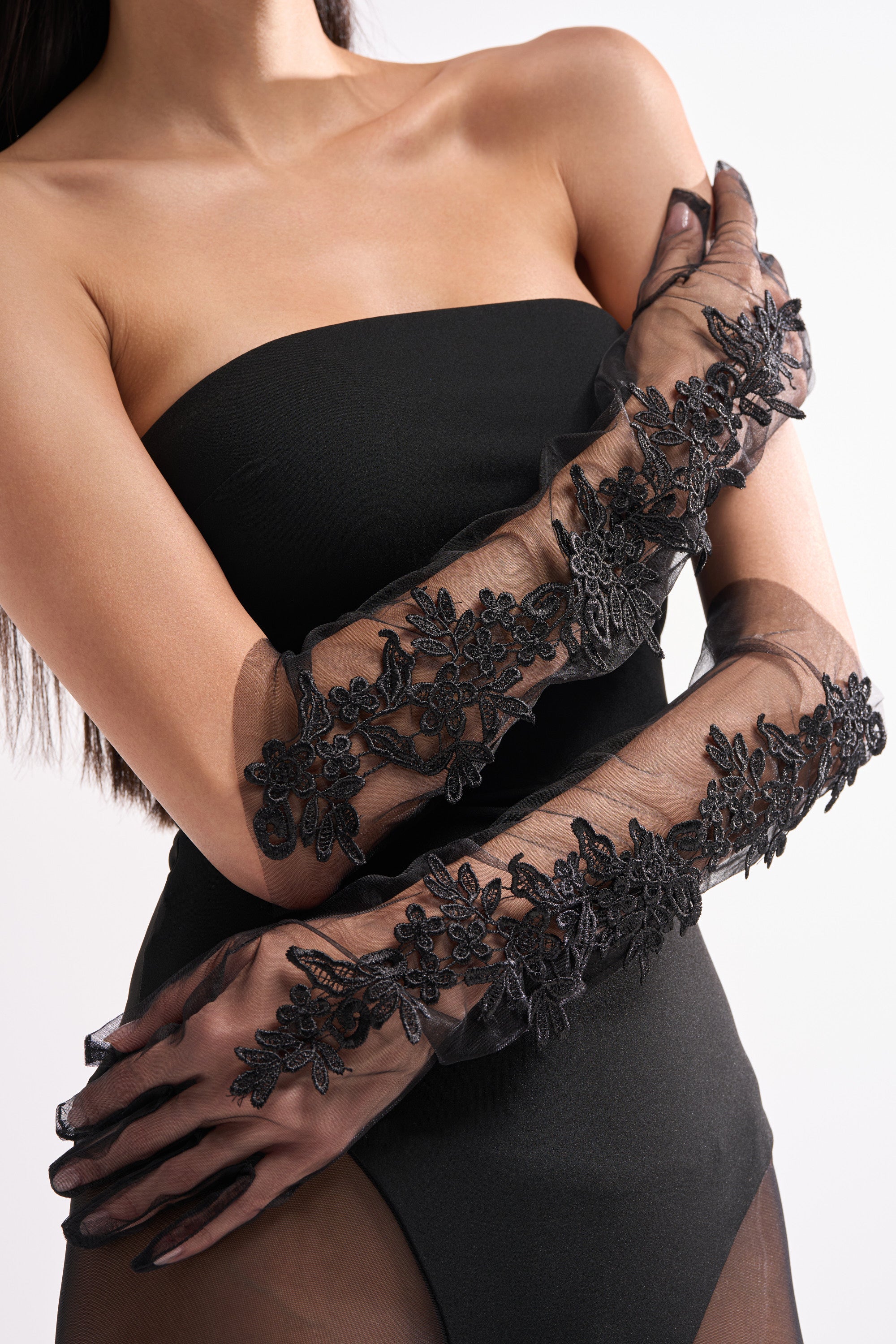 A woman crosses her arms in front of her chest, wearing the FLORA MESH GLOVE IN BLACK—sheer, floral-embroidered gloves—paired with a strapless black dress against a plain light background.