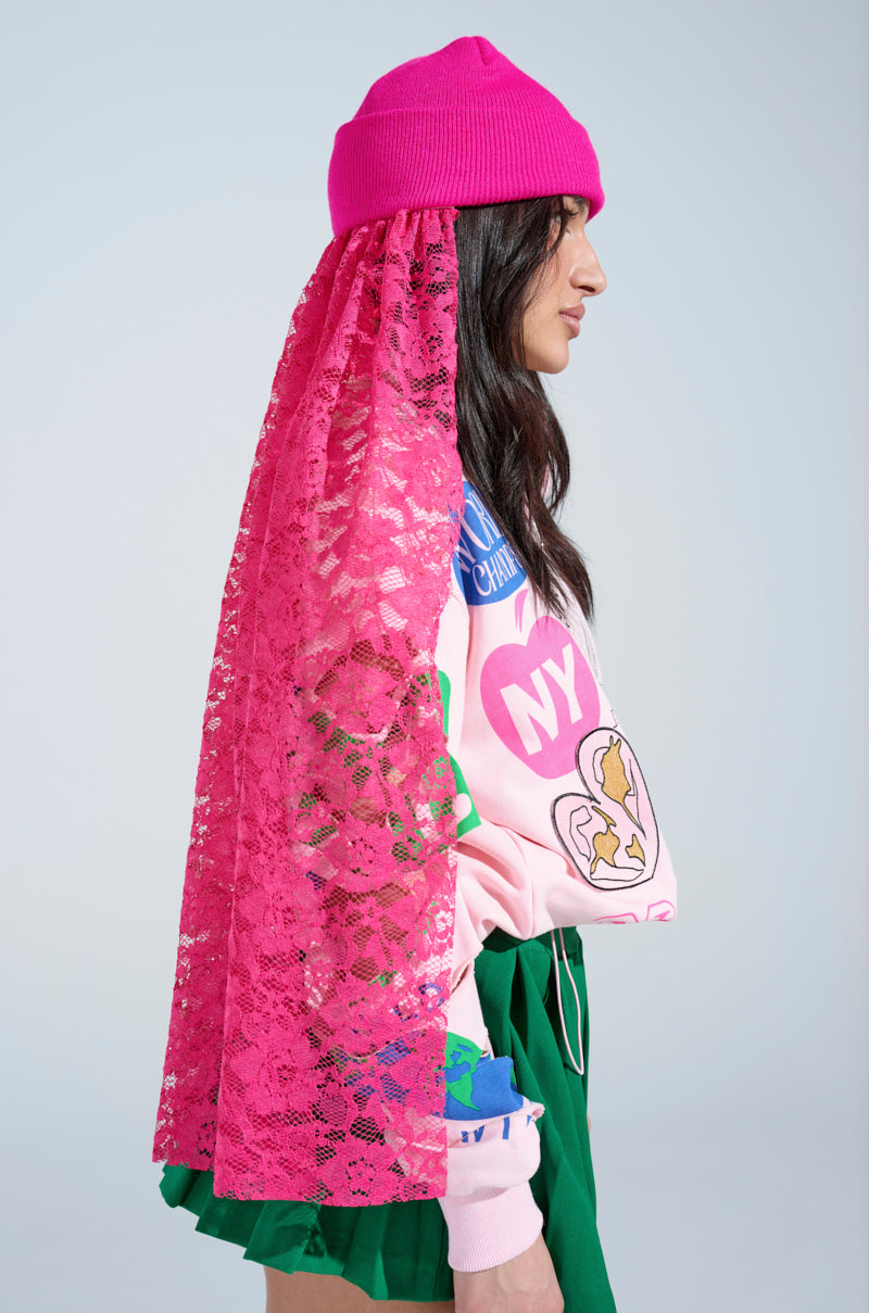 Wearing an IN THE CUT LACE VEIL BEANIE in vibrant hot pink, paired with a colorful graphic sweater and a green skirt, a person poses in profile against a light blue backdrop, gazing to the right.