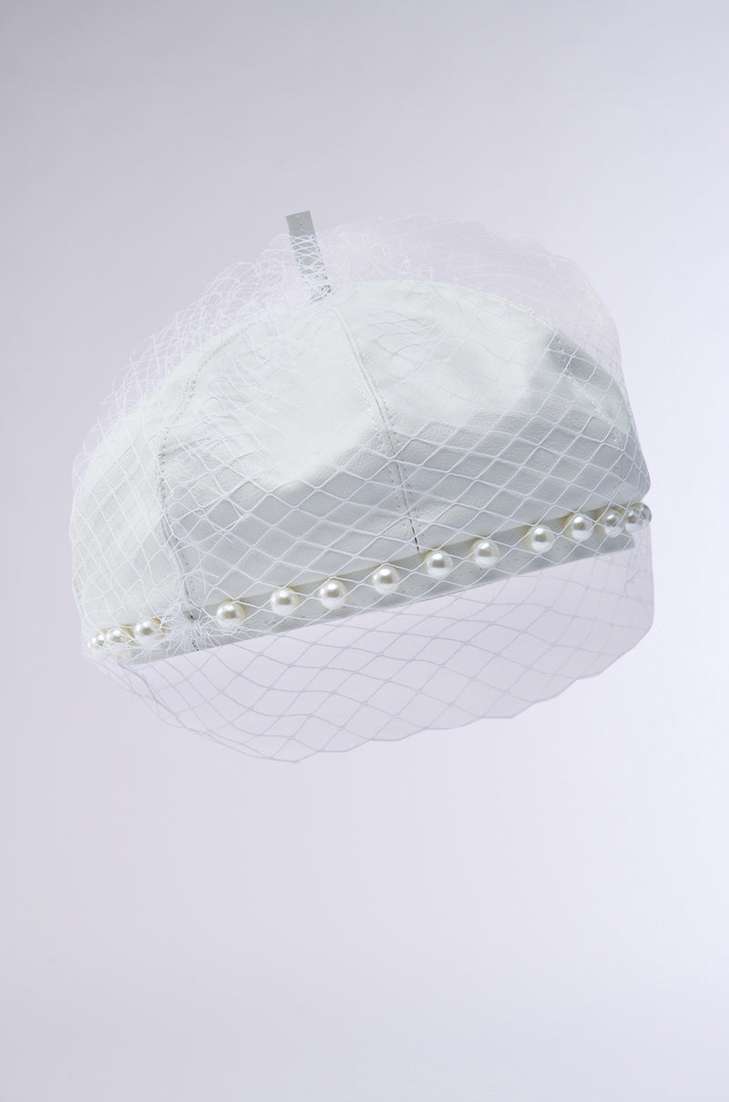 The EDITORIAL BERET, made of vegan leather in white, features a pearl-adorned trim and delicate netting. Its tonal mesh veil elegantly drapes over the front against a light, neutral background, adding sophistication.
