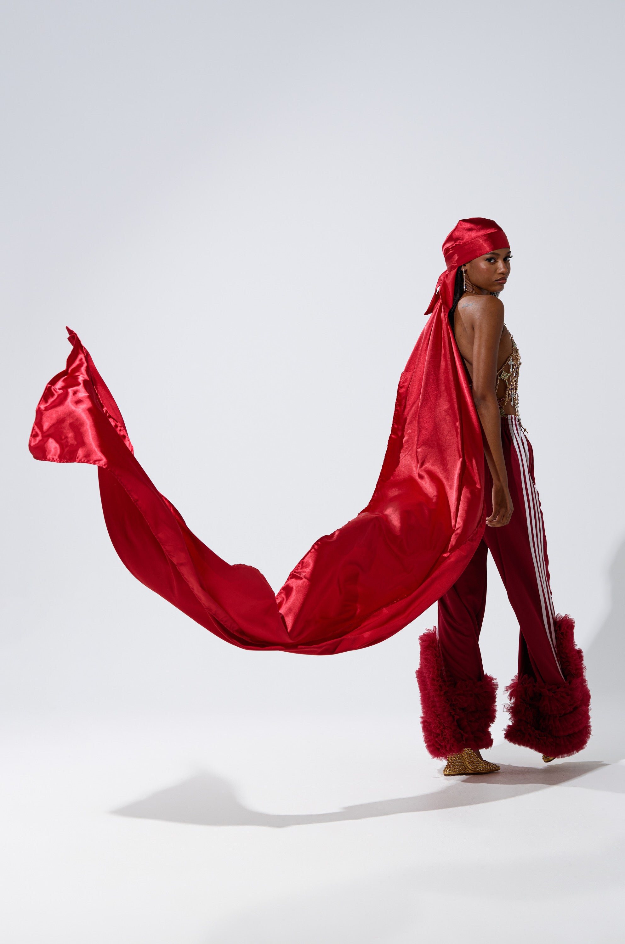 A person poses confidently against a plain white background, wearing THE MOMENT EXTENDED SATIN DURAG styled floor-length, along with a long red cape, striped pants, and furry red leg warmers.