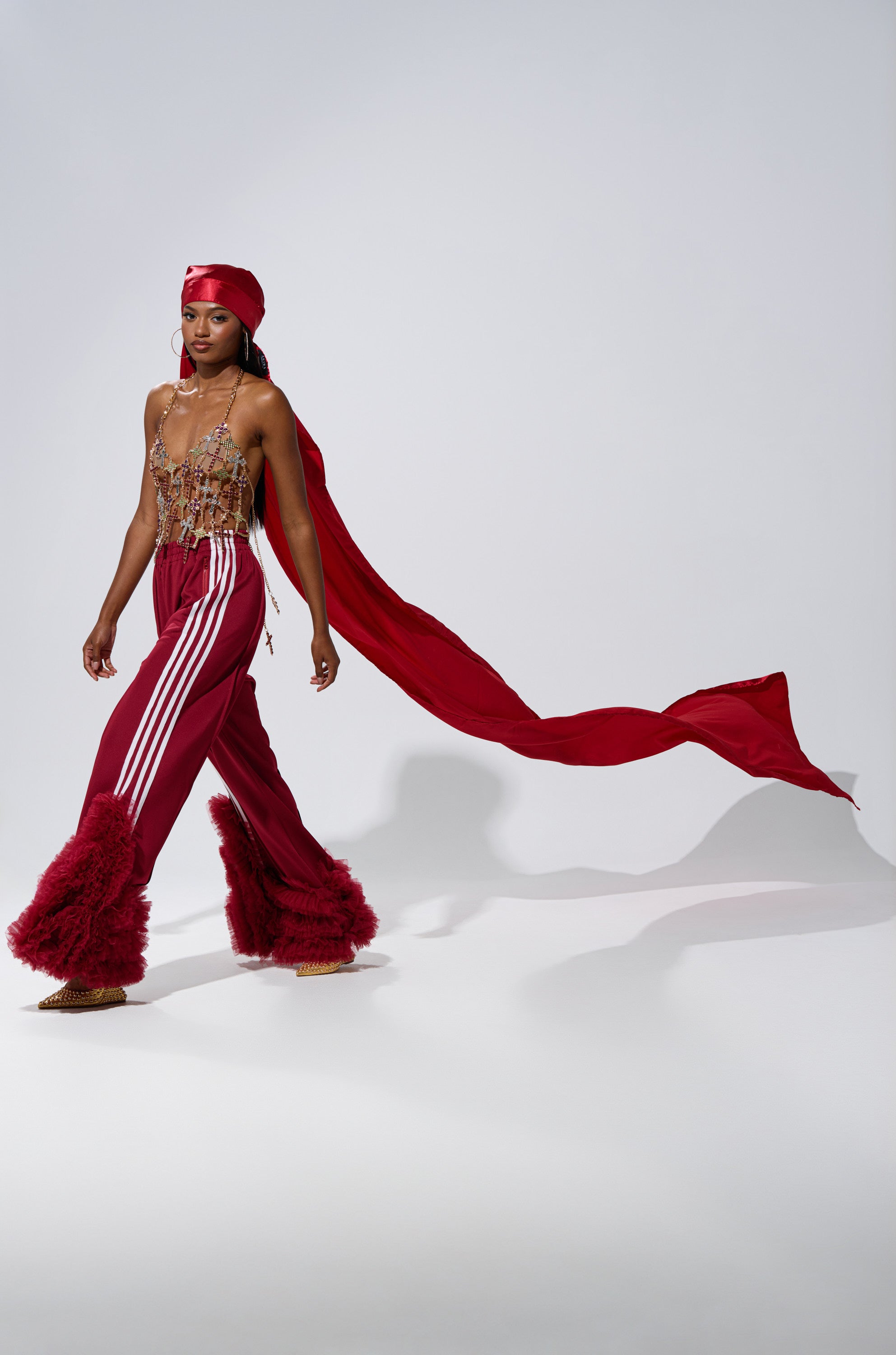 Wearing THE MOMENT EXTENDED SATIN DURAG, a person in red flared pants with white stripes and a jeweled top walks confidently against a plain light background.