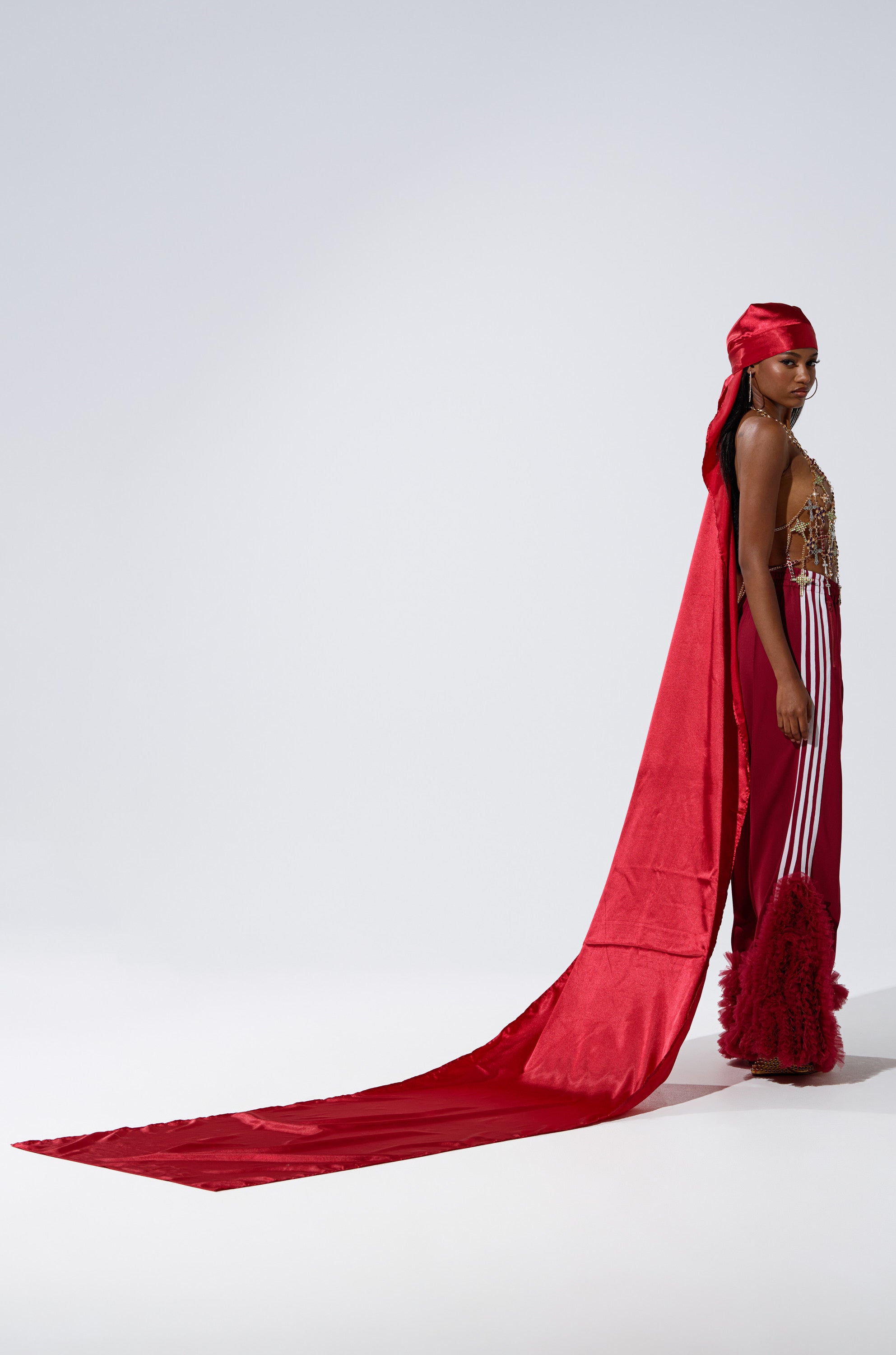A person stands confidently in THE MOMENT EXTENDED SATIN DURAG, paired with a long red satin cape, an embellished top, and red pants with fluffy textured hems against a plain light background.