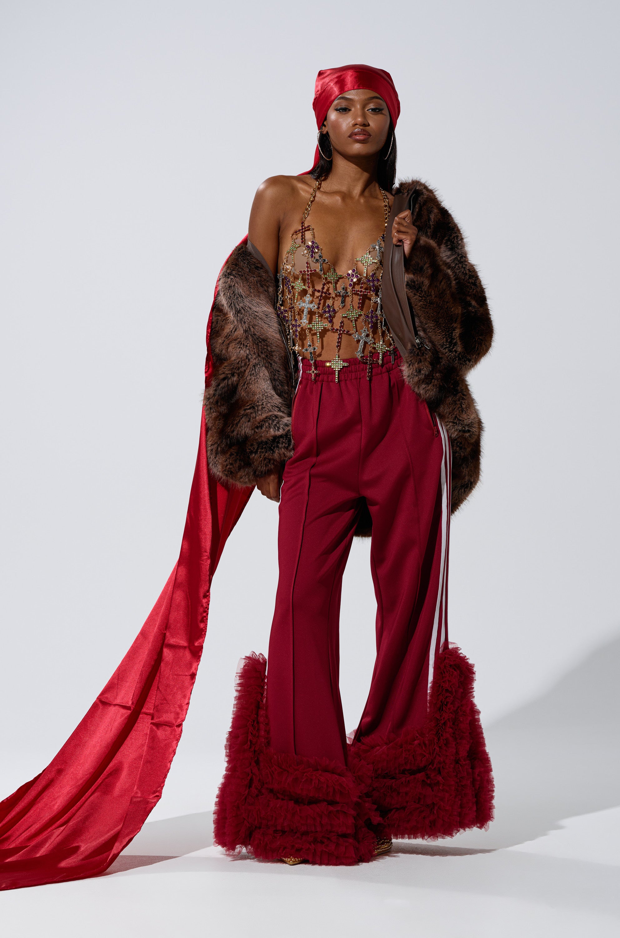 A woman poses in THE MOMENT EXTENDED SATIN DURAG, a beaded top, faux fur jacket, red wide-leg pants with ruffled hems, and a flowing red sash against a plain light background.