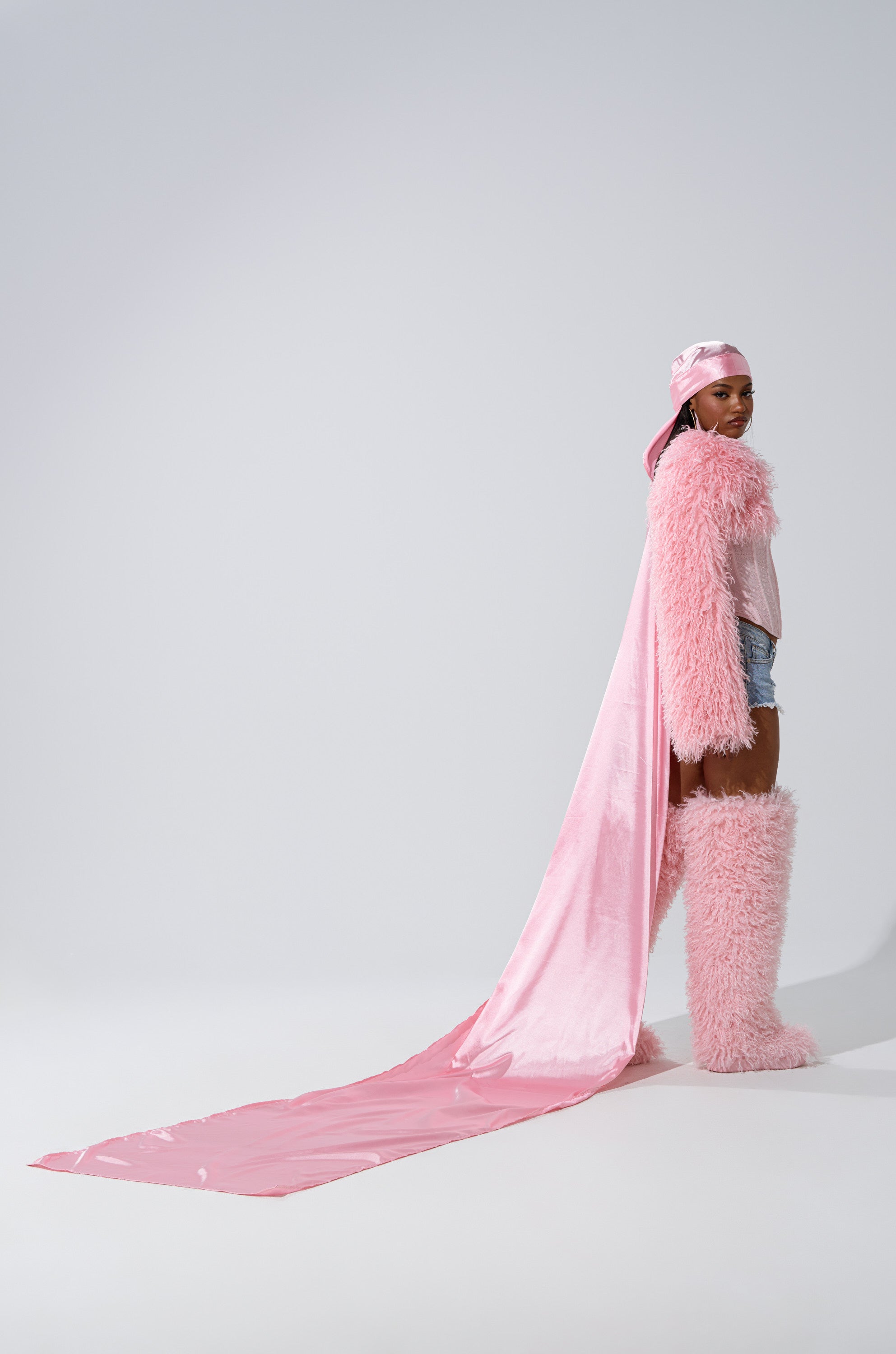 A person stands sideways against a plain light background wearing denim shorts, a pink furry jacket and matching boots, and the MAKE MY ENTRANCE EXTENDED DURAG with a long flowing pink satin cape.