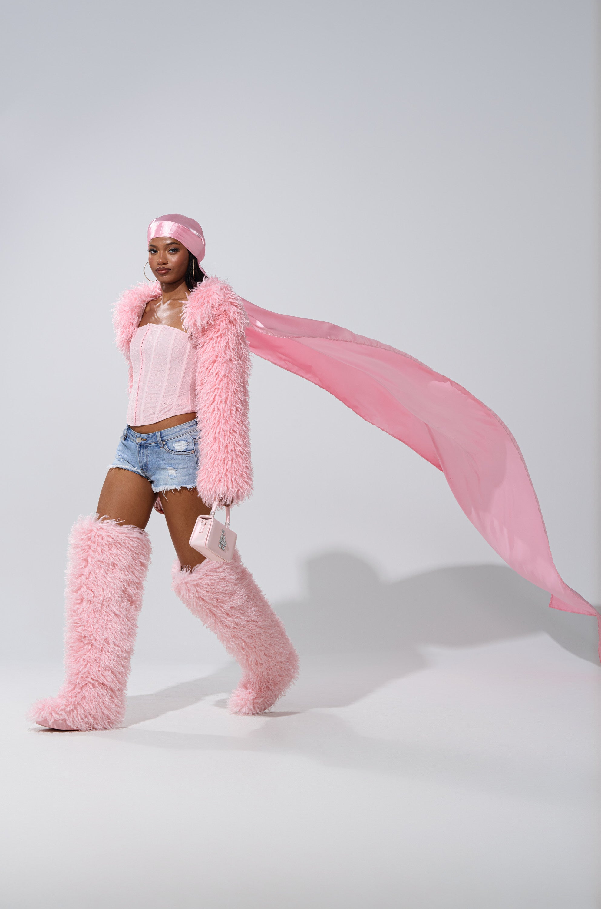 Rocking the MAKE MY ENTRANCE EXTENDED DURAG, a person in a fluffy pink coat, furry boots, denim shorts, pink corset, long scarf, and holding a small pink purse walks confidently against a plain background.