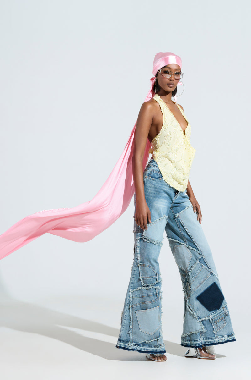 A person poses confidently in a light yellow halter top and patchwork flared jeans, with the "MAKE MY ENTRANCE EXTENDED DURAG" flowing behind them like a silhouette, accessorized with large hoop earrings and clear-framed glasses.