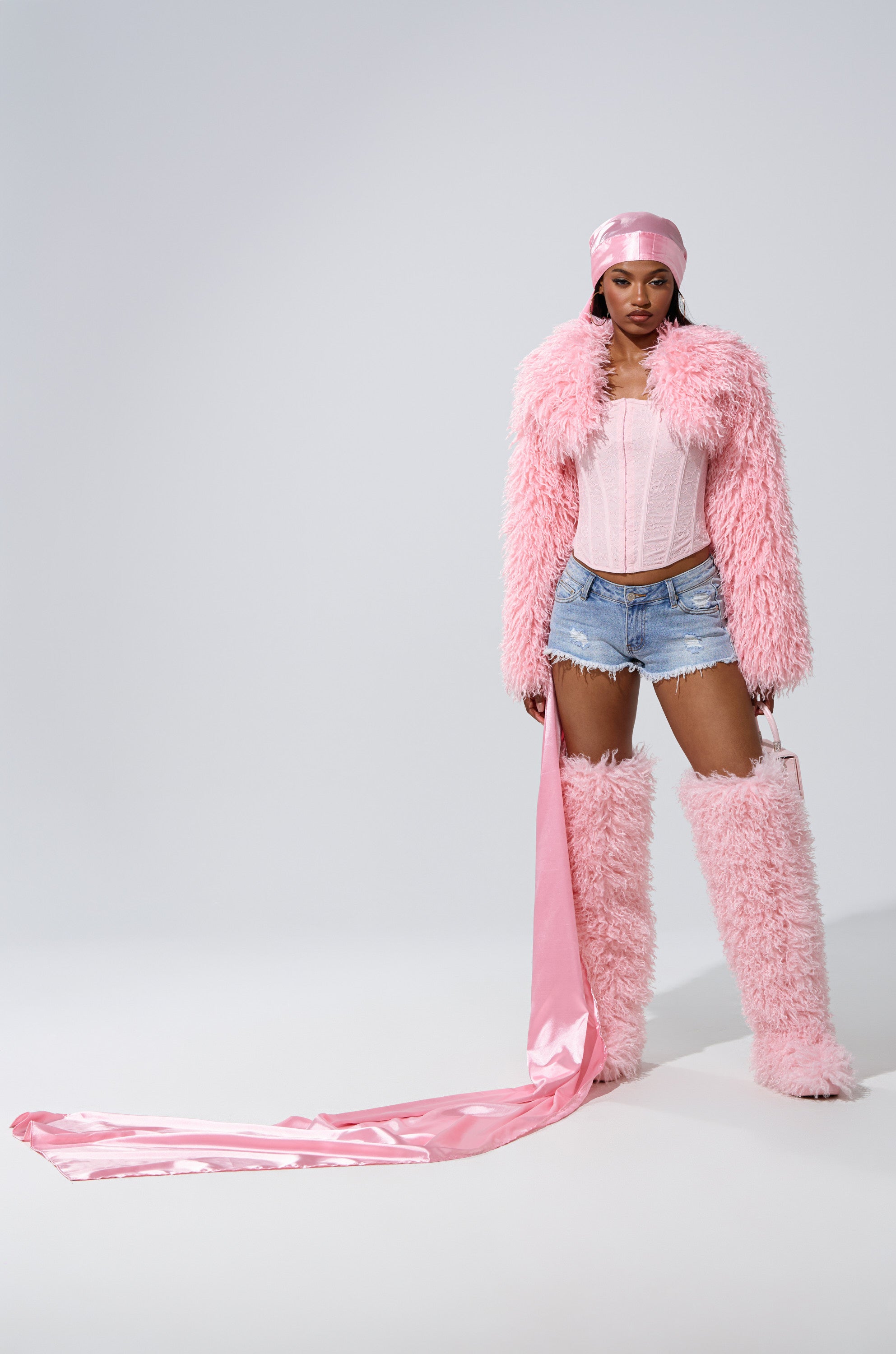 A woman stands against a plain light background in a pink fuzzy coat, matching tall boots, pink corset top, ripped denim shorts, and a long satin sash, her look finished with the MAKE MY ENTRANCE EXTENDED DURAG.