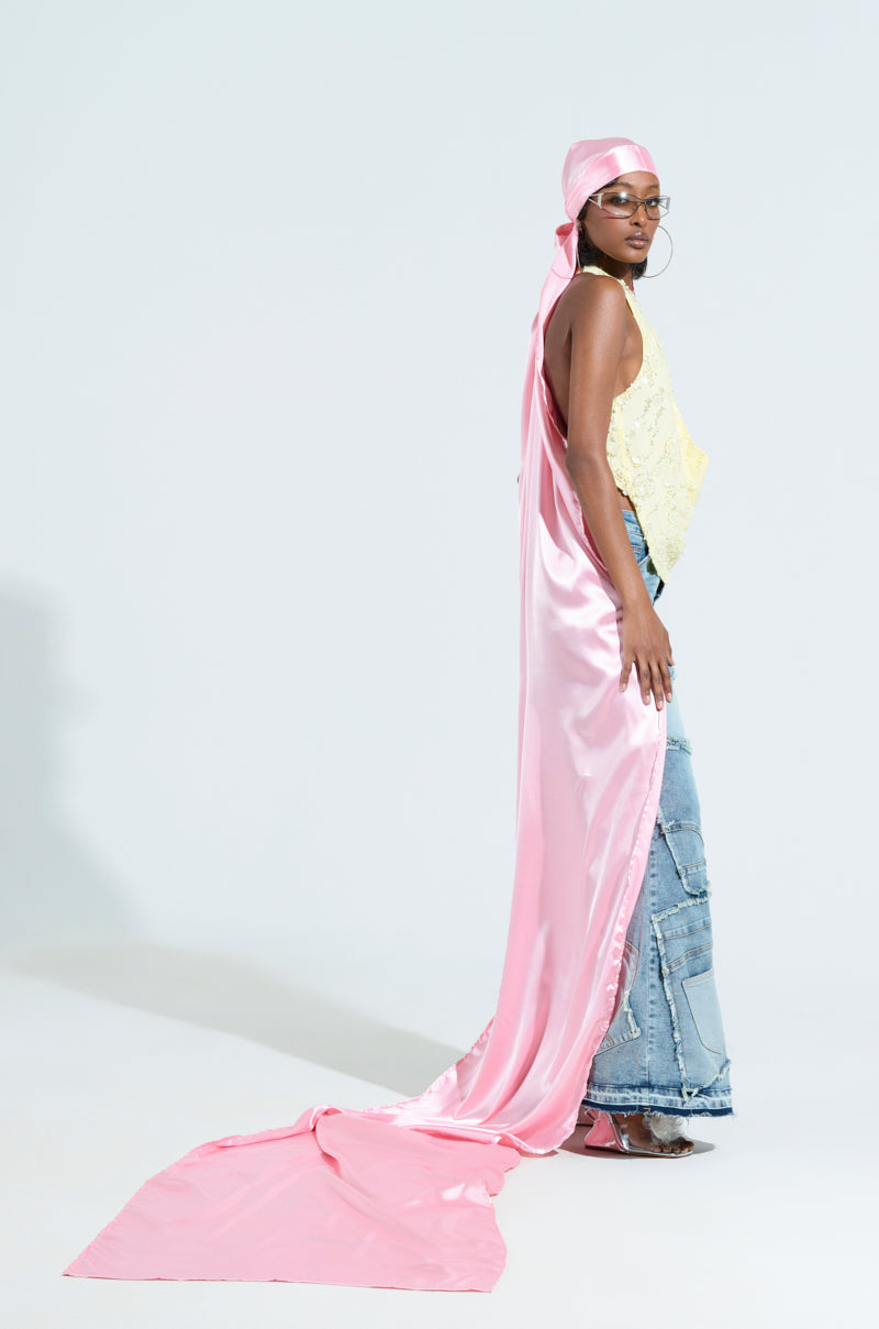 Wearing the MAKE MY ENTRANCE EXTENDED DURAG and a long, flowing pink satin headscarf, a person confidently poses in a yellow lace top and blue patchwork jeans. With glasses on, they strike a stylish pose against a plain white backdrop.