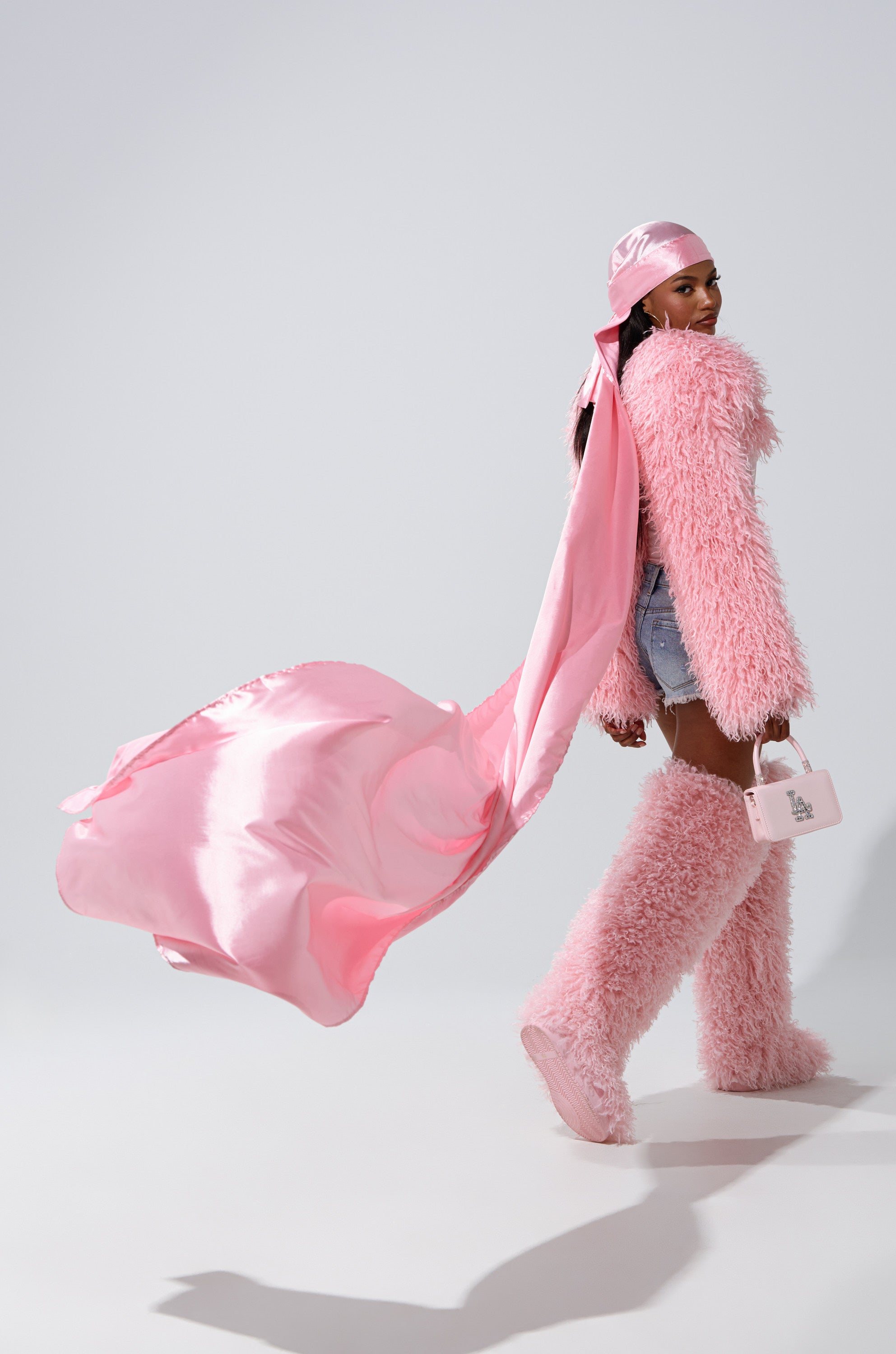 A woman wearing the MAKE MY ENTRANCE EXTENDED DURAG in floor-length pink satin, a fluffy pink coat, matching furry boots, and denim shorts holds a small pink purse while looking over her shoulder against a plain background.