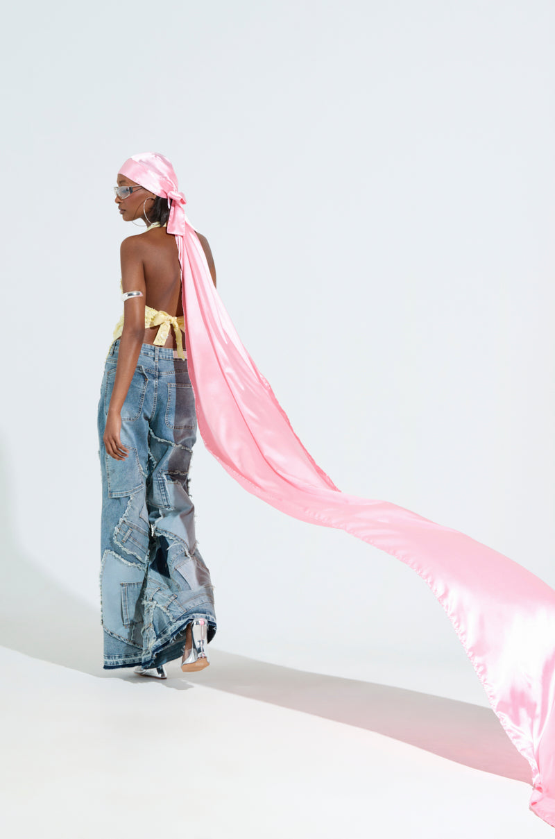 They are wearing a long, flowing Make My Entrance Extended Durag in pink and sunglasses, paired with a strapless top and patchwork denim pants, set against a white background with the silky scarf dramatically extending behind them.