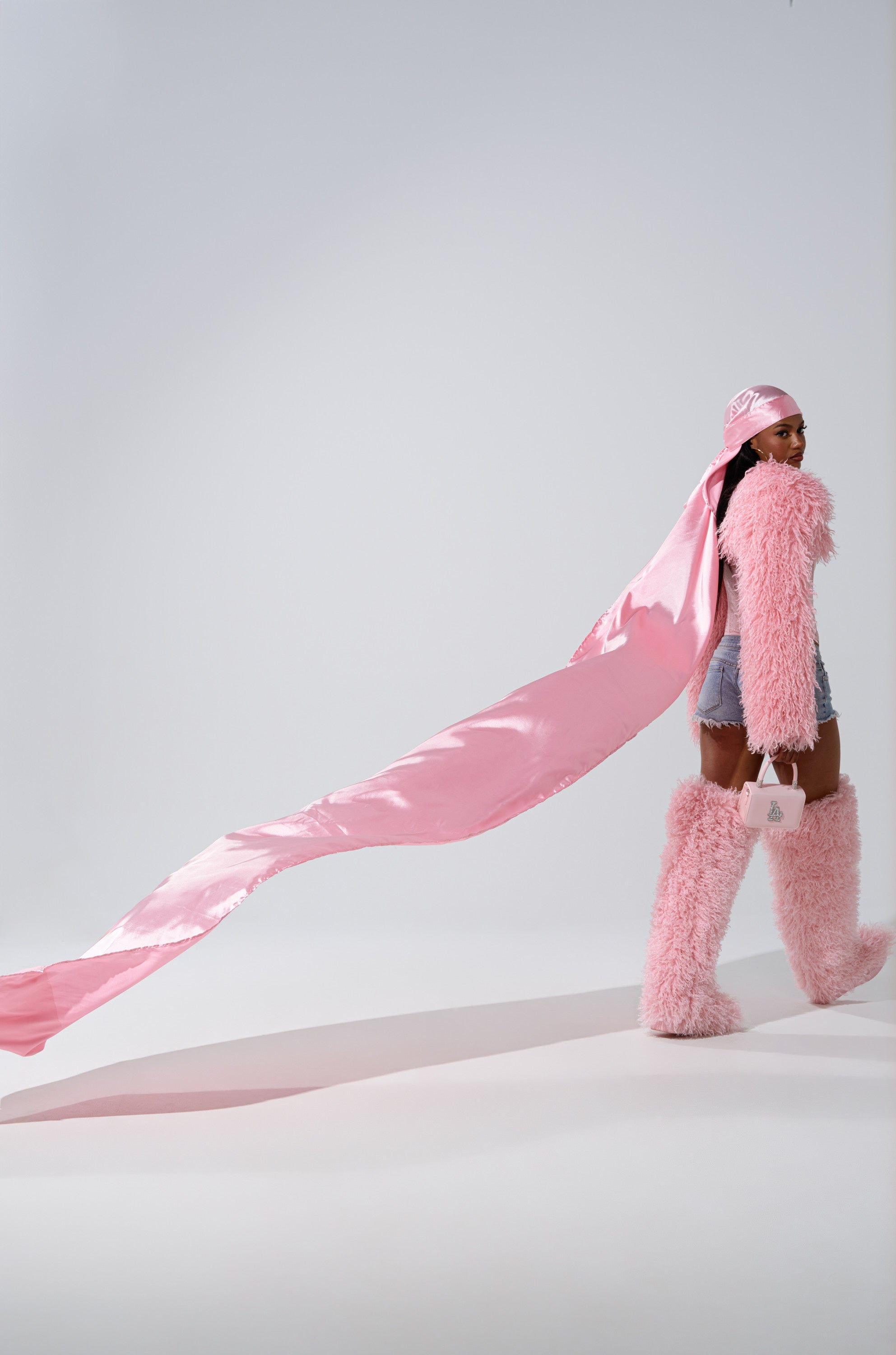 Wearing the MAKE MY ENTRANCE EXTENDED DURAG in floor-length pink satin, along with a matching headscarf, fluffy pink boots, a furry pink jacket, and holding a small pink purse, they pose against a plain white background.