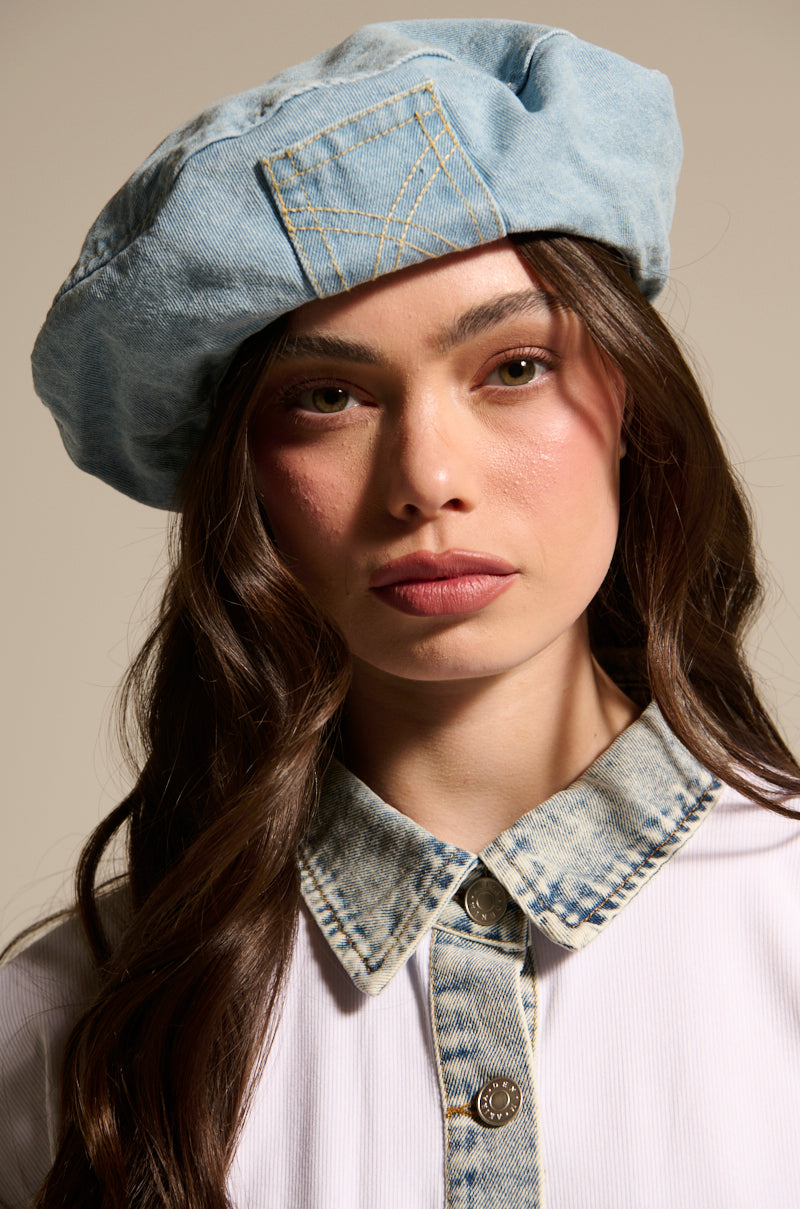 A woman with long brown hair wears the IT GURL DENIM BERET from AKIRA Label and a white shirt with a bleached denim collar, looking directly at the camera with a neutral expression against a plain background.