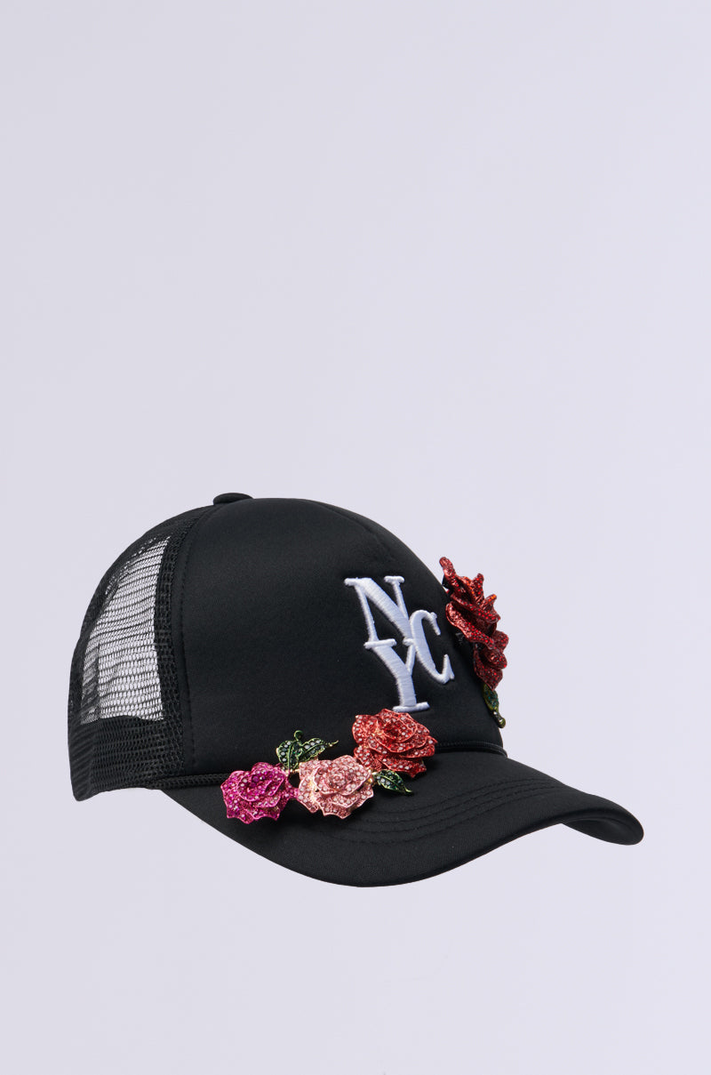 The BIG CITY BROOCH TRUCKER HAT is a black trucker cap with "NYC" embroidered in white on the front. It features colorful sequined floral designs on the brim and side for added flair, and has a black mesh back panel for a classic finish.