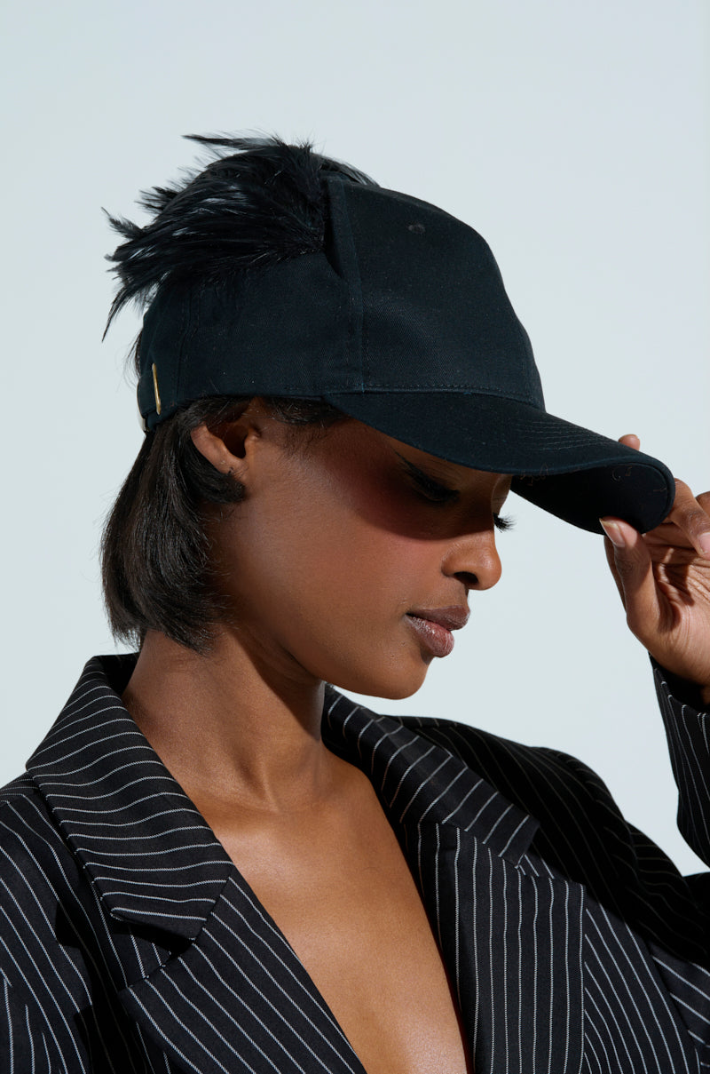 A person in a pinstriped blazer adjusts the brim of their BRB FLYING AWAY HAT, featuring an adjustable back strap and side feather details, against a plain, light background.