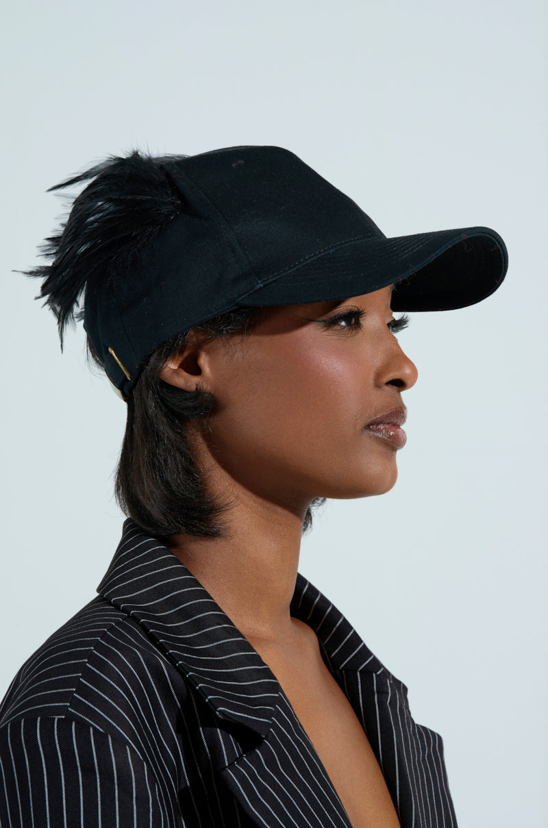 A person in profile sports a BRB FLYING AWAY HAT—a black baseball cap featuring a feather detail and adjustable back strap—stylishly paired with a black pinstripe blazer, their short hair accentuated by the light background.