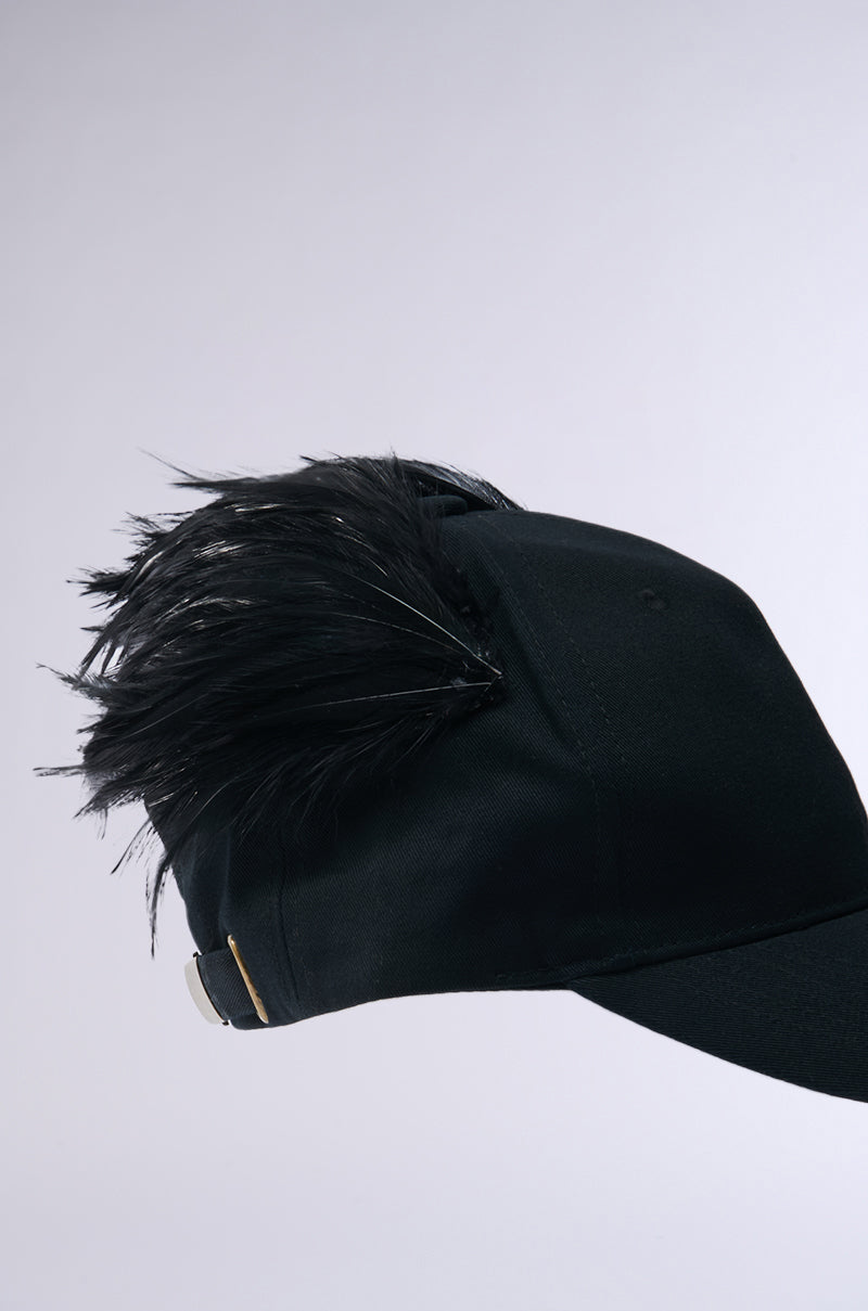 The BRB FLYING AWAY HAT is a distinctive black baseball cap with elegant black feathers atop and at the back. It's crafted from cotton with a sleek metal clasp on its adjustable strap, featured against a light, neutral background.