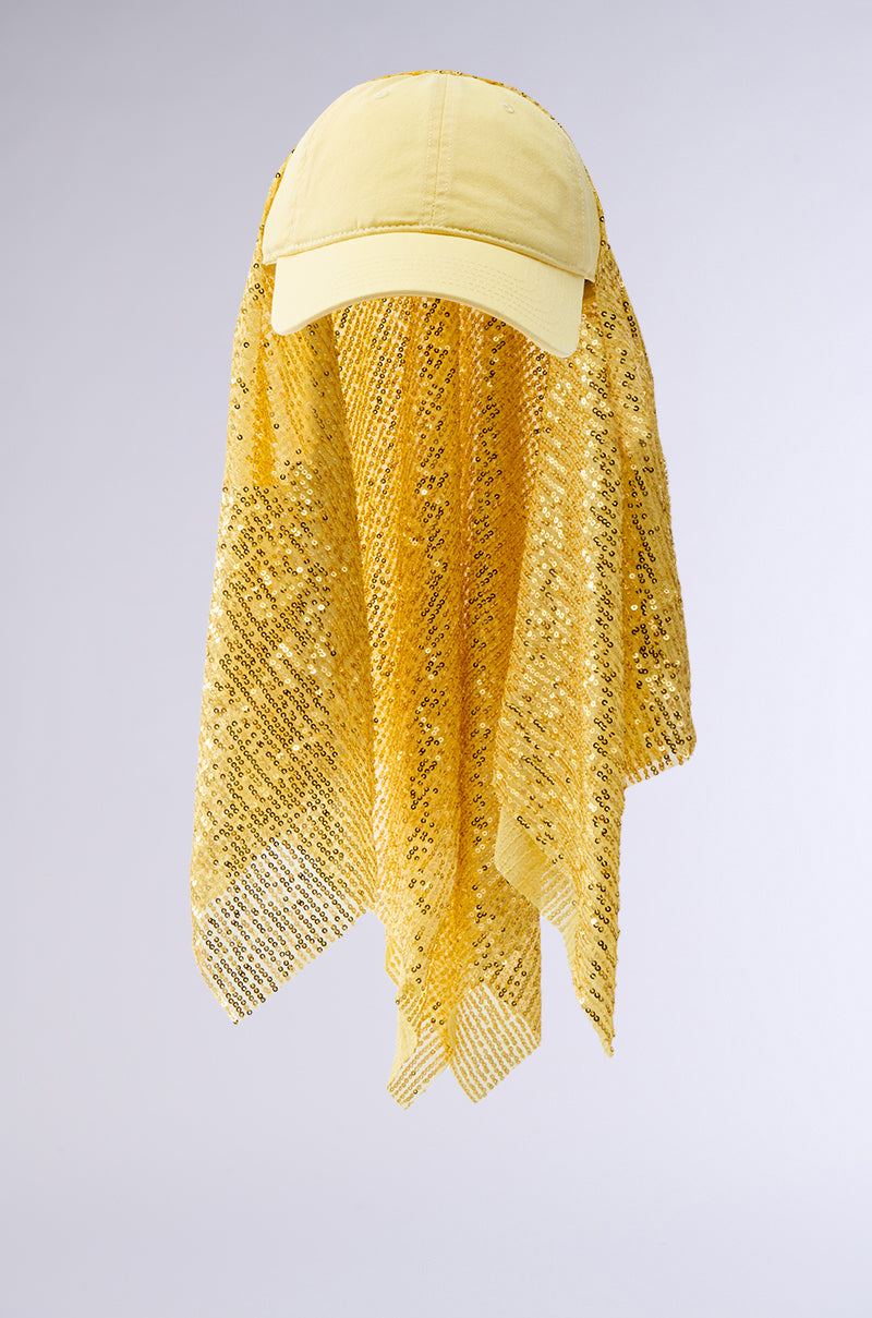 The IN NYC SEQUIN SCARF HAT is a yellow hat with tonal sequin embellishments, a long sparkly mesh drape at the back, and an adjustable buckled strap. Shown on a plain light gray background.