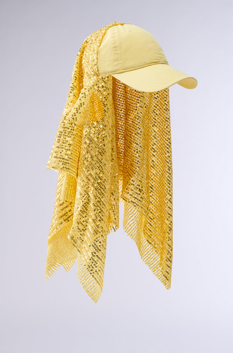 The IN NYC SEQUIN SCARF HAT features a yellow baseball cap with a long, sheer yellow lace scarf draping like a veil and an adjustable buckled strap at the back, shown against a plain light gray background.