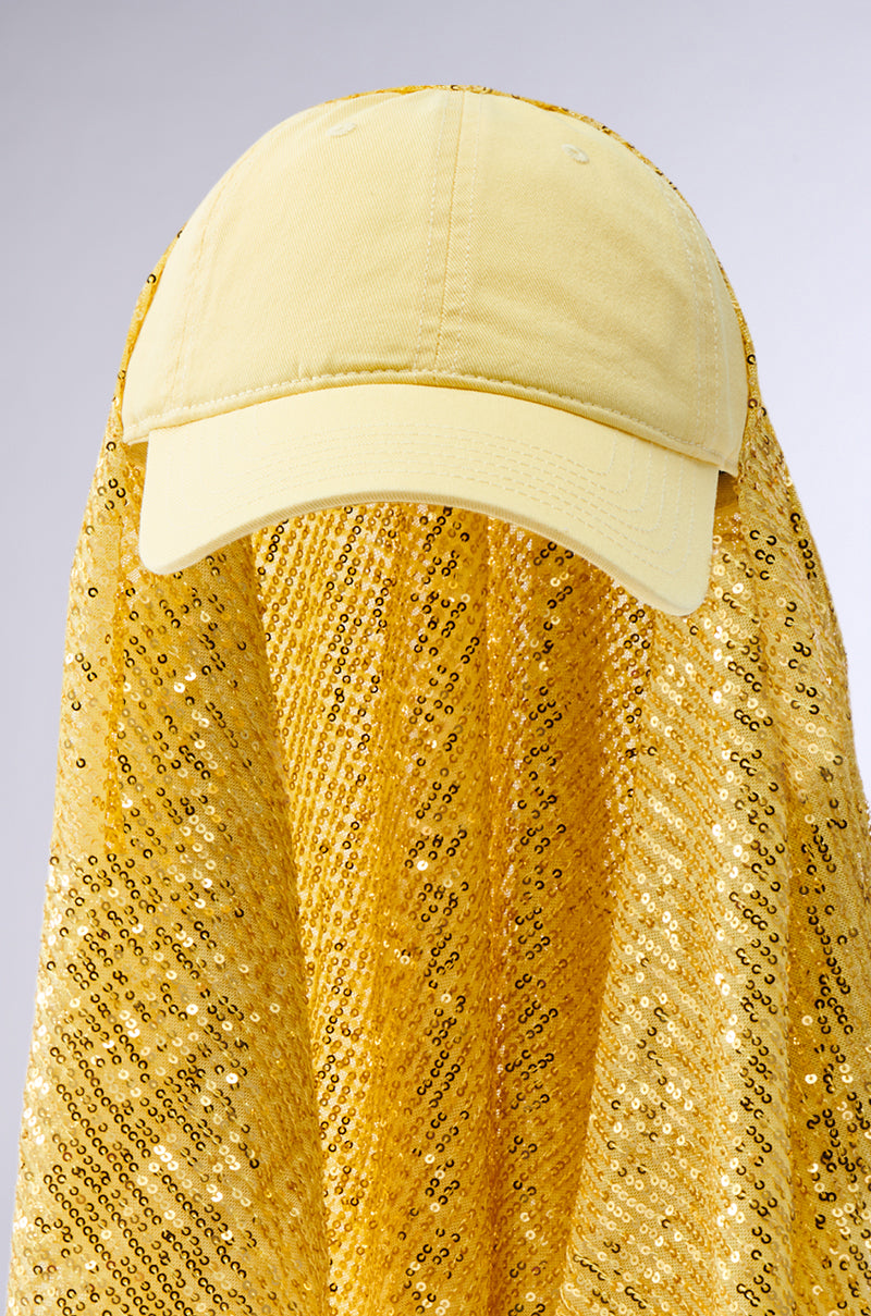 The IN NYC SEQUIN SCARF HAT, a yellow baseball cap with sequins and an adjustable buckle strap, covers the entire body and face in matching tonal embellishments against a neutral gray background.