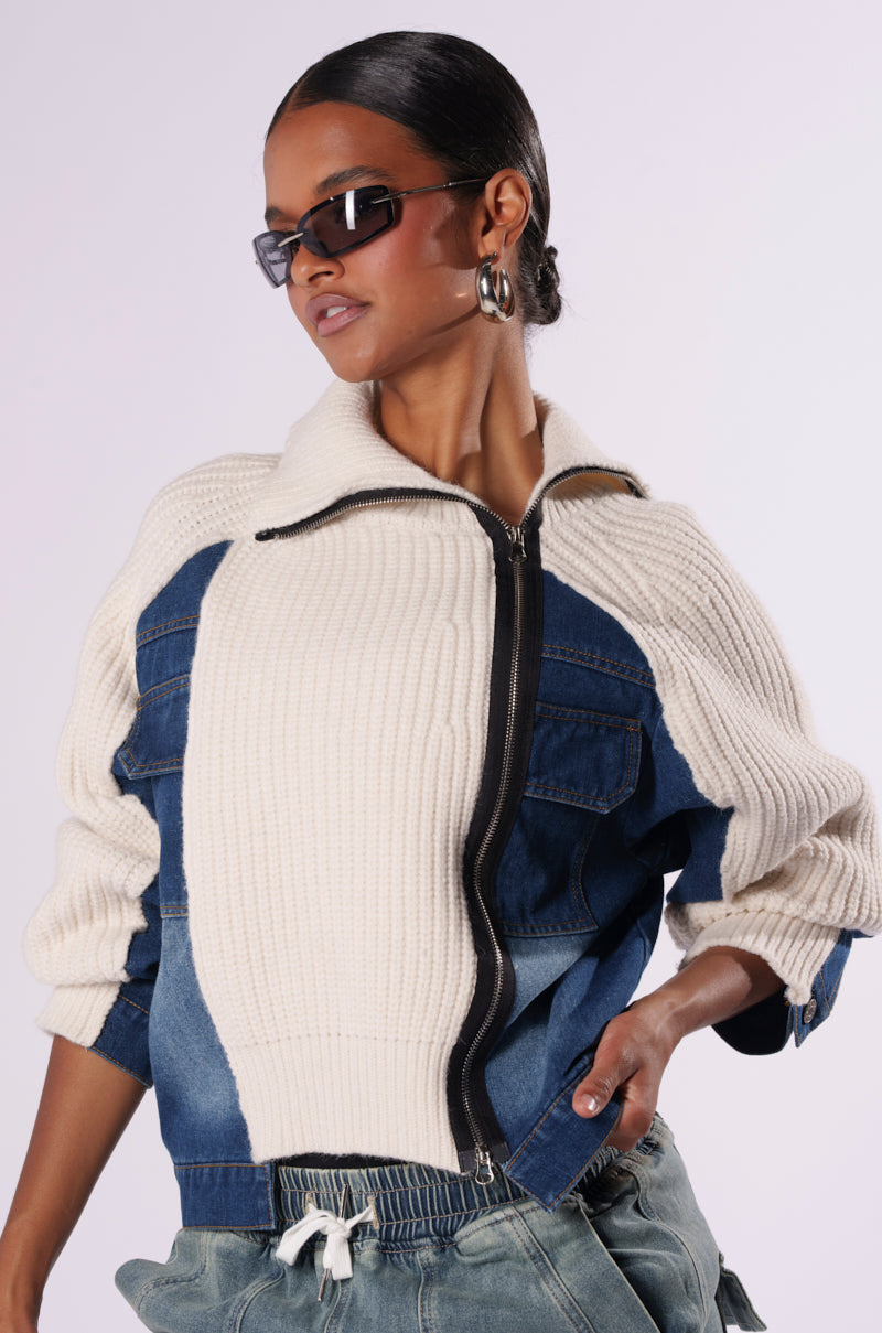 A person is confidently posing in the SOMETHING ABOUT YOU SWEATER, a stylish combination of a woven knit base paired with dark wash denim sections. The top, which includes a prominent front zipper and large pockets, pairs perfectly with their sunglasses and large hoop earrings. Their hair is styled back, completing the chic look.